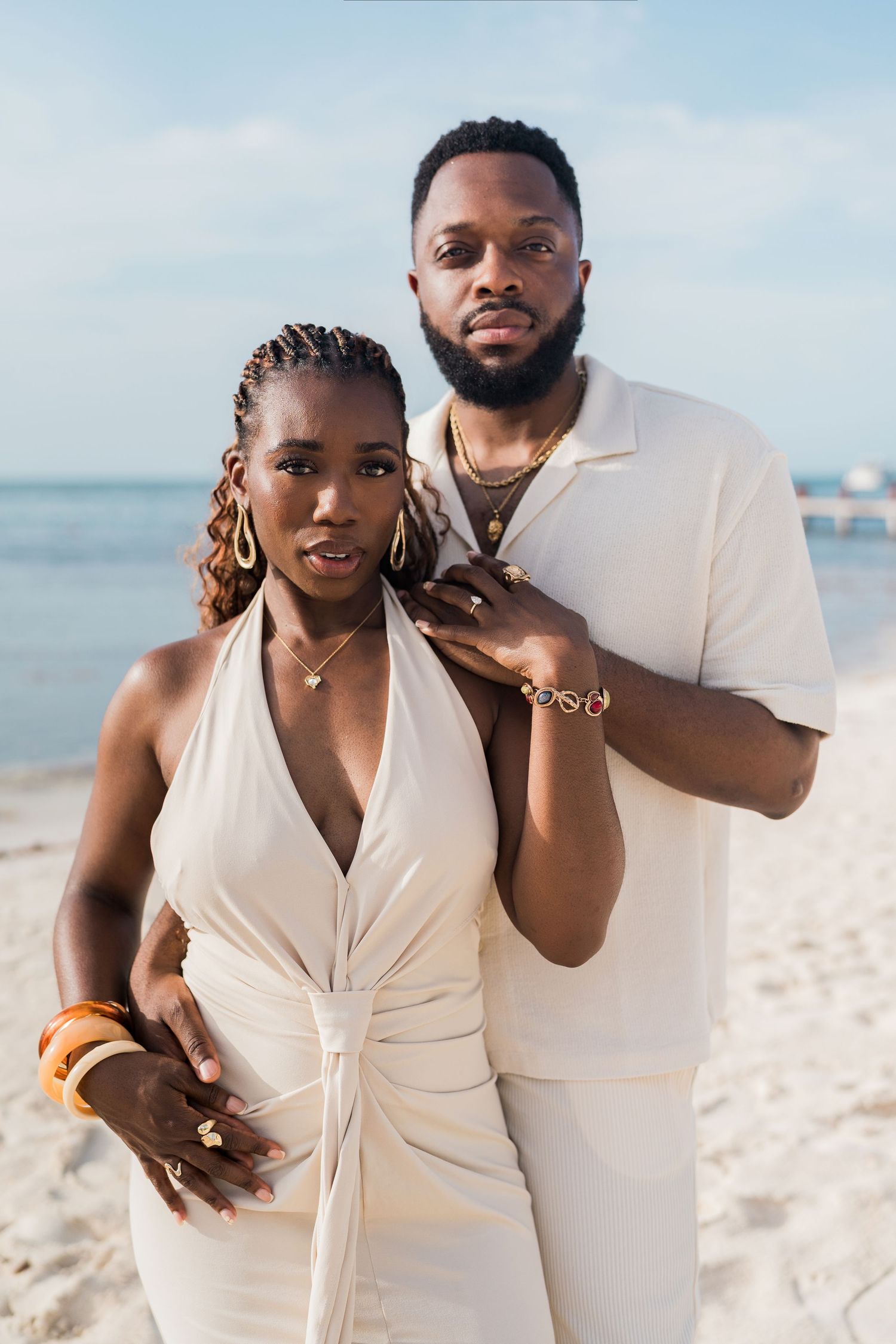 Elegant Black couple posing on a sandy beach in matching white outfits and gold jewelry.