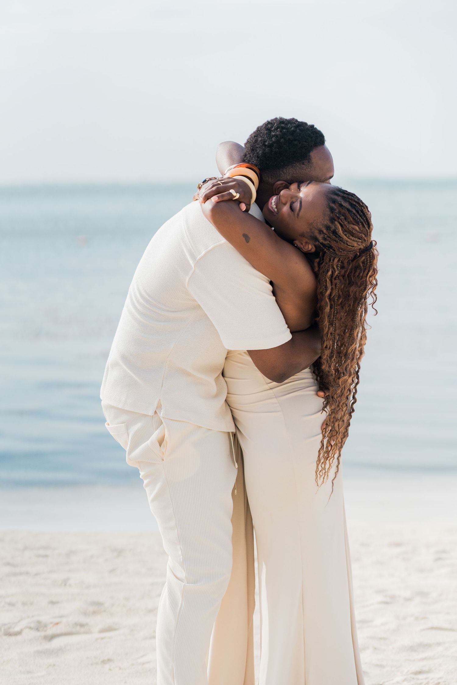 A couple in white attire shares a romantic kiss on a sandy beach with calm ocean waters in the background.