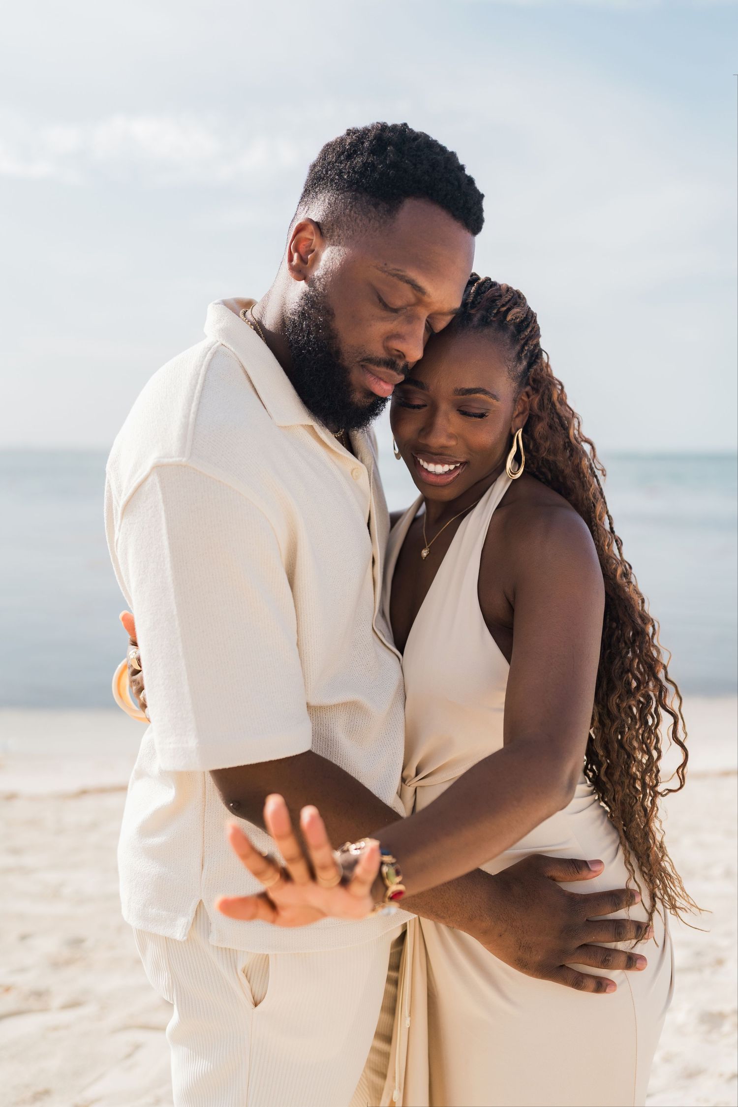 Happy couple embraces on a sunny beach, woman smiling while wearing cream dress and gold hoop earrings.
