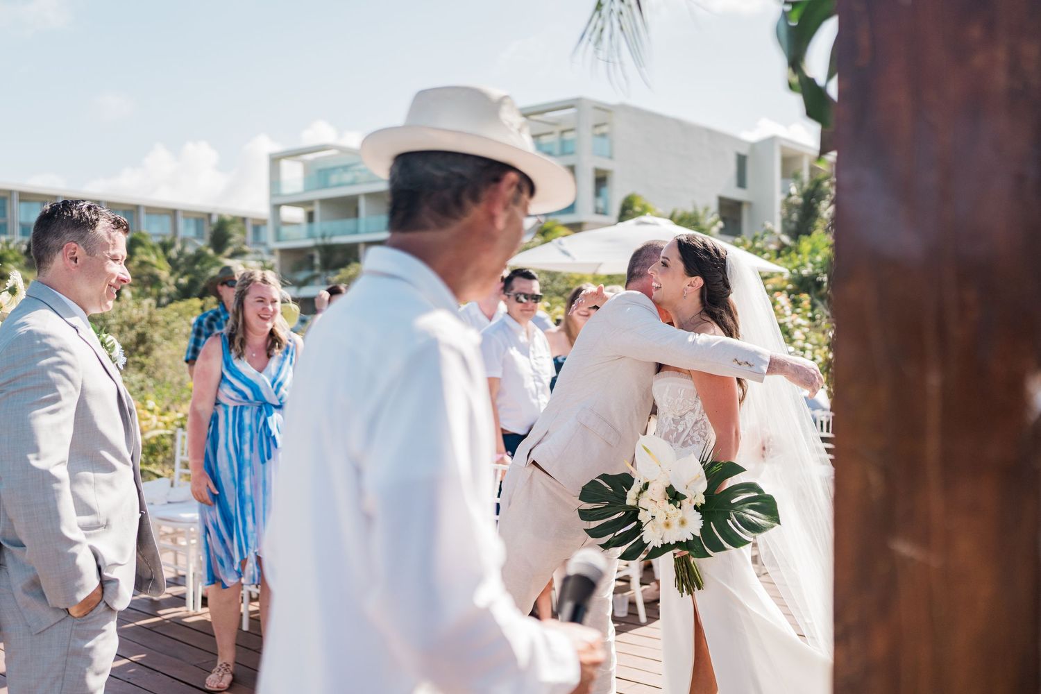 Bride in white gown hugs guest at outdoor beach wedding ceremony with tropical decor and modern resort backdrop.