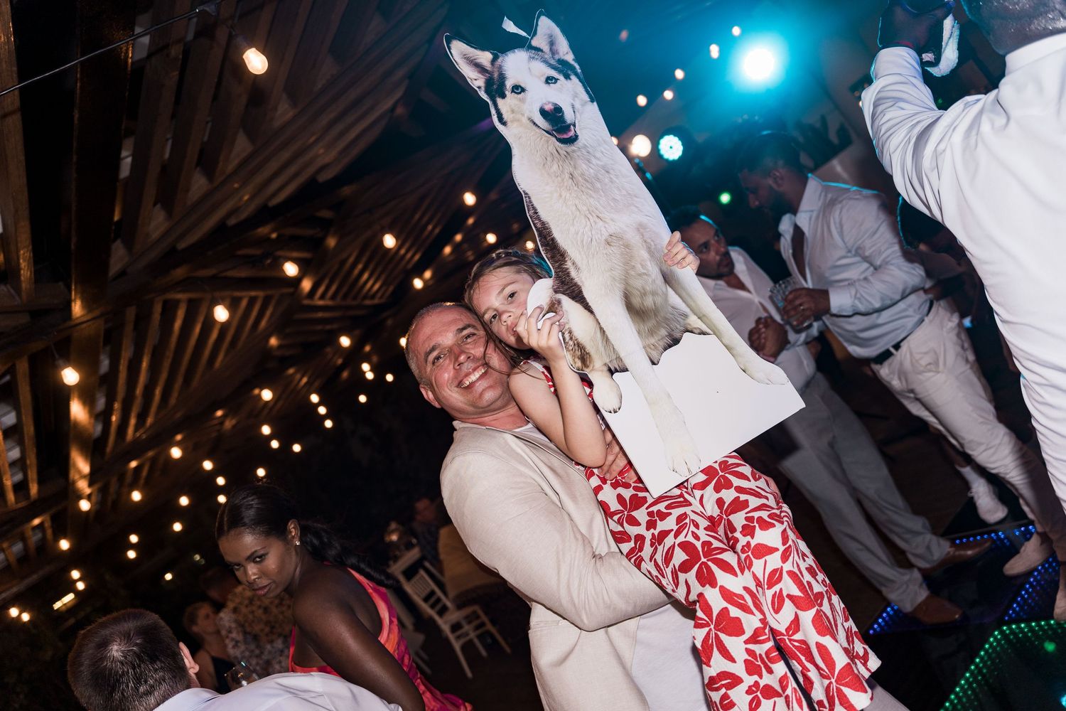 Joyful wedding guest holds a large cutout of a Husky dog at a festive barn reception with string lights.