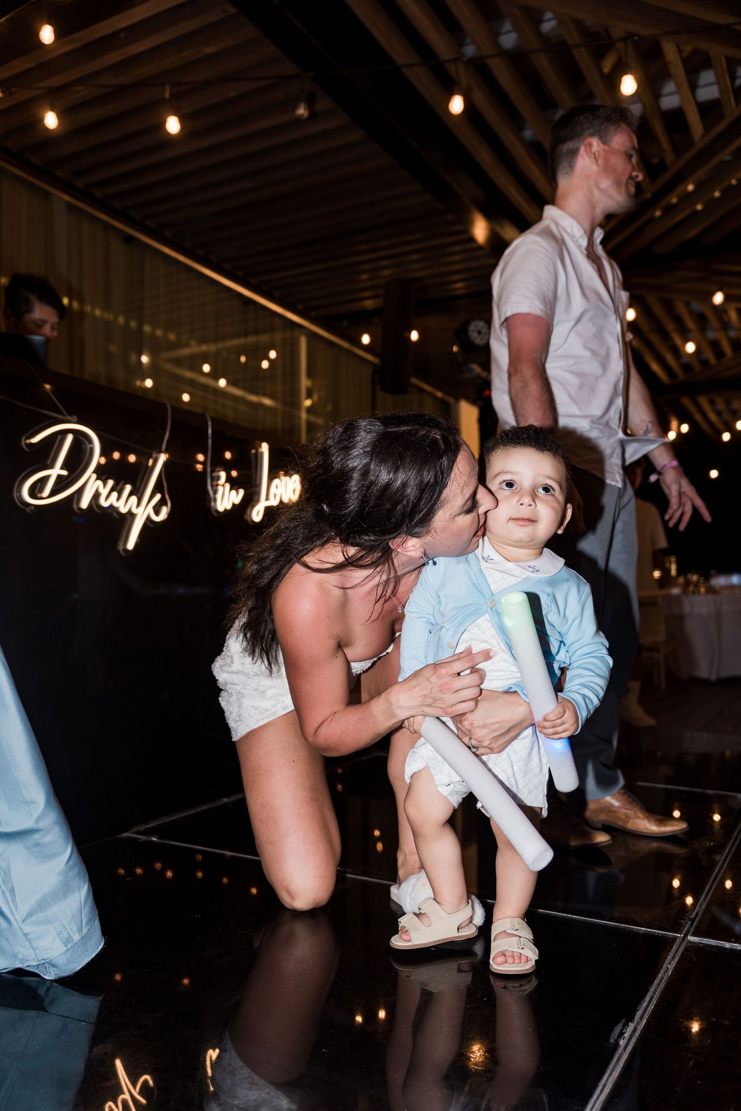 A bride kneels on a dance floor kissing a toddler at a wedding reception with romantic neon sign lighting.
