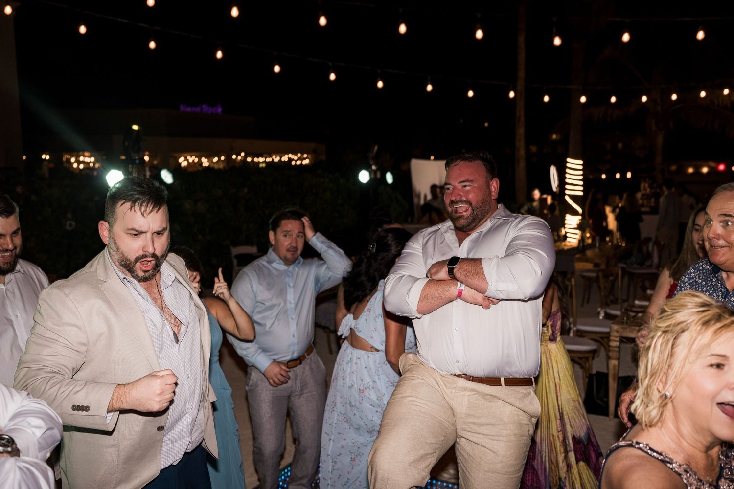 Men in white shirts dancing energetically at an outdoor evening wedding reception with string lights.