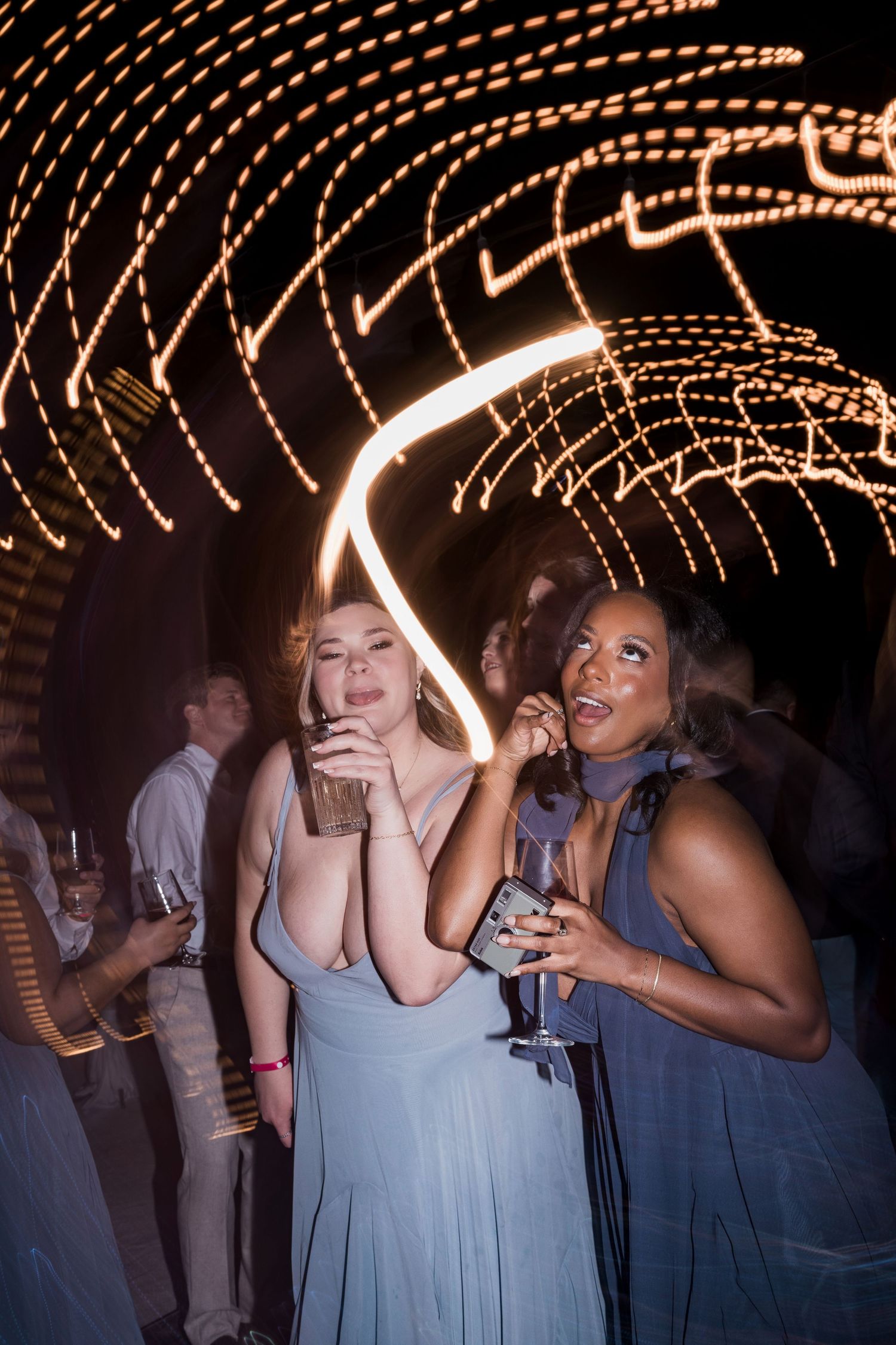 Two women in blue gowns dancing at a glamorous night event with glowing light trails in the background.