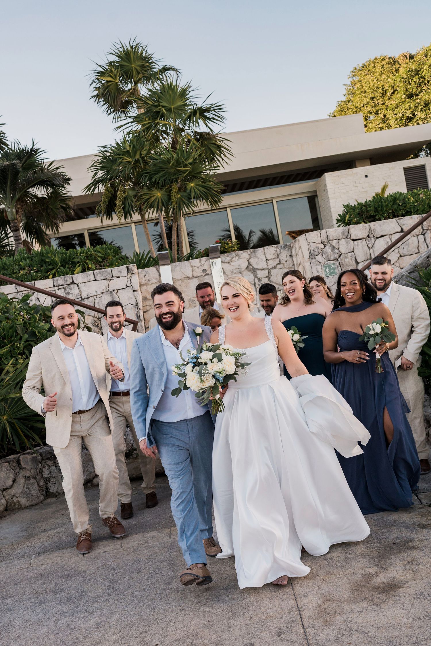 Wedding party walks together outdoors, bride in white gown with bouquet, groom in blue suit, groomsmen and bridesmaids in navy.