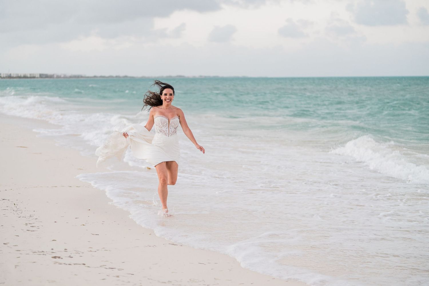 Bride in white dress running along a sandy beach with ocean waves, hair flowing in the wind on a cloudy day.