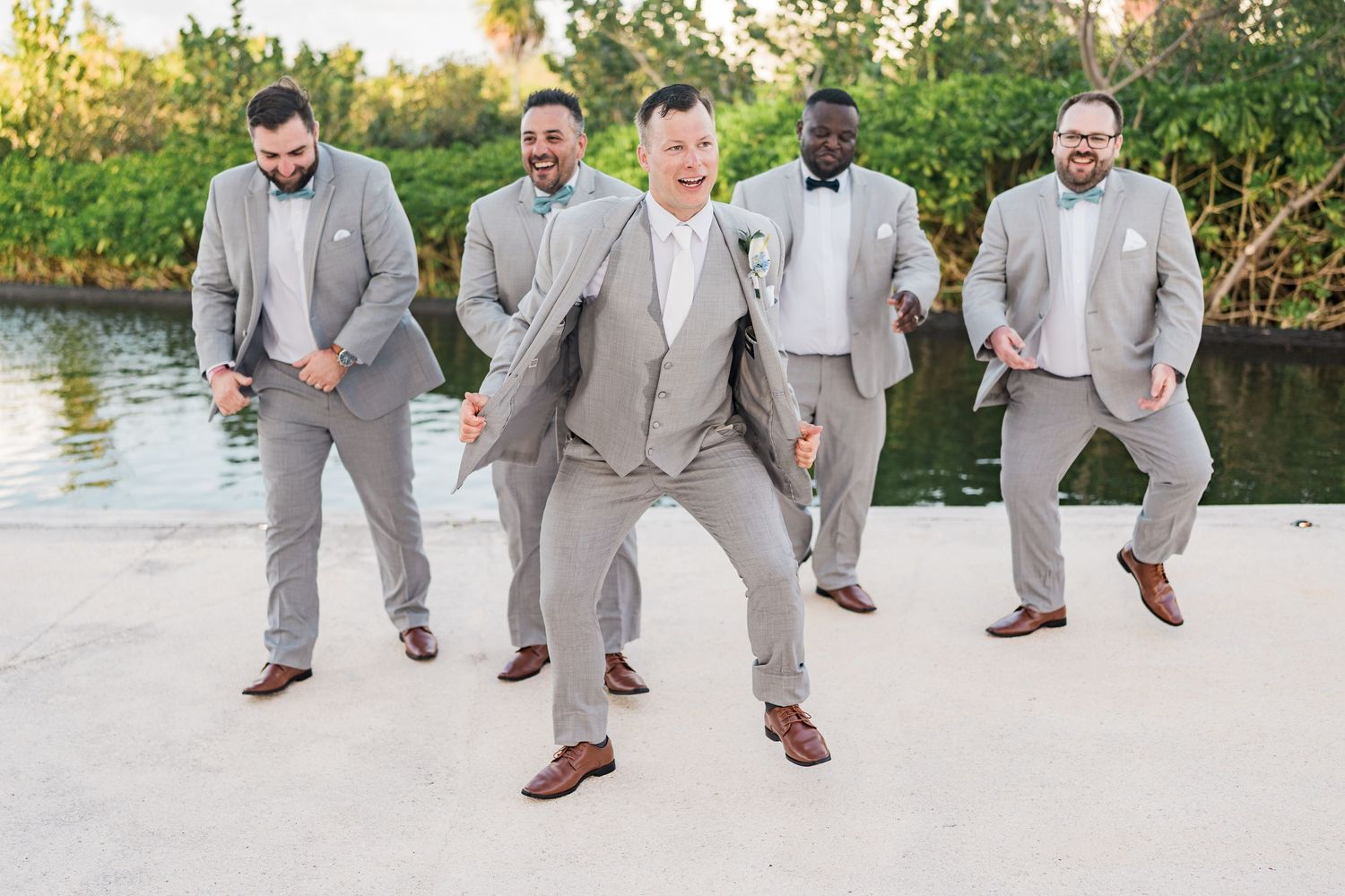 Five groomsmen in gray suits dancing and posing outdoors near a waterfront at a wedding celebration.