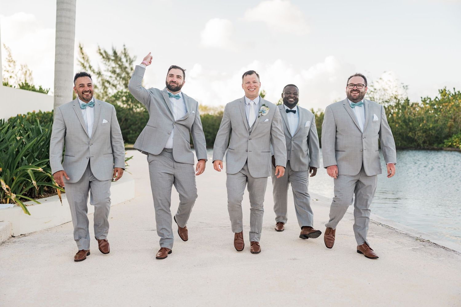 Five groomsmen in light gray suits with bow ties walk confidently outdoors near a waterfront at a tropical wedding.