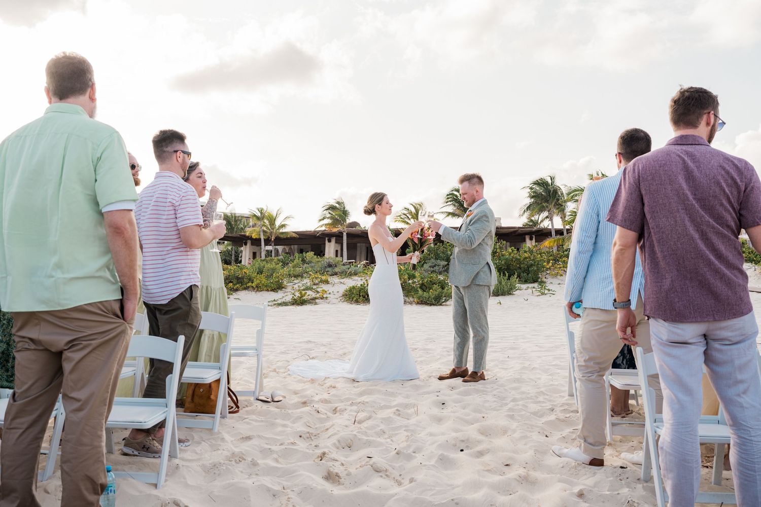 Couple exchanges vows at a romantic beach wedding ceremony with guests standing on white sand near tropical resort.