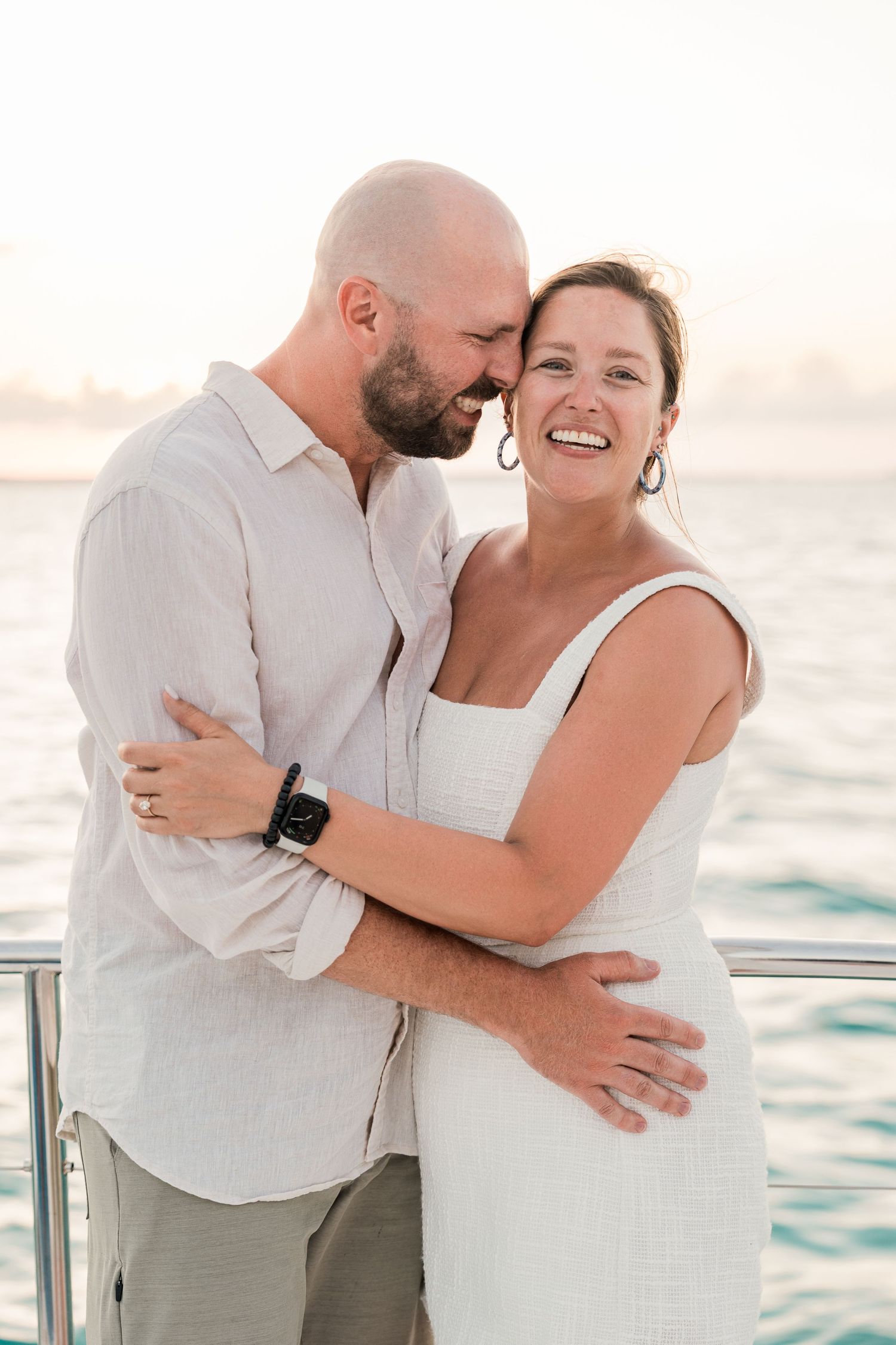 Happy couple embracing on a yacht at sunset, woman in white dress smiling, man in linen shirt, ocean backdrop.