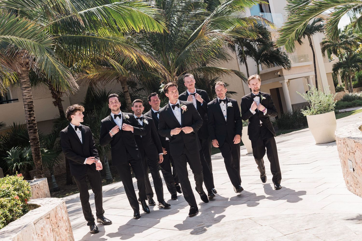 Groomsmen in black tuxedos and bow ties walk together outdoors near palm trees at a tropical wedding venue.