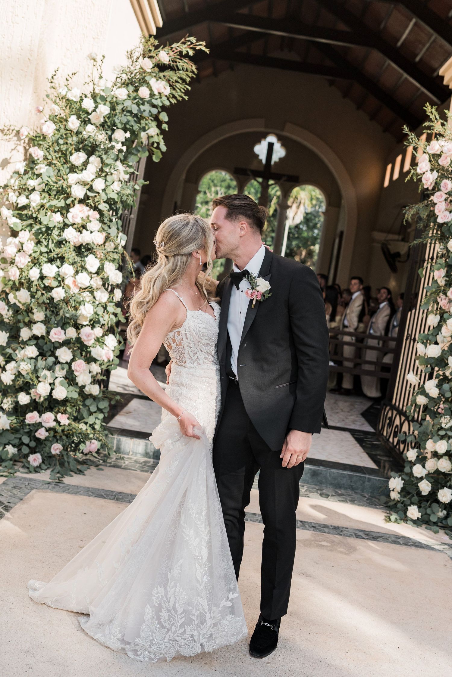 Bride and groom share a kiss outside a church adorned with lush white floral arrangements on their wedding day.