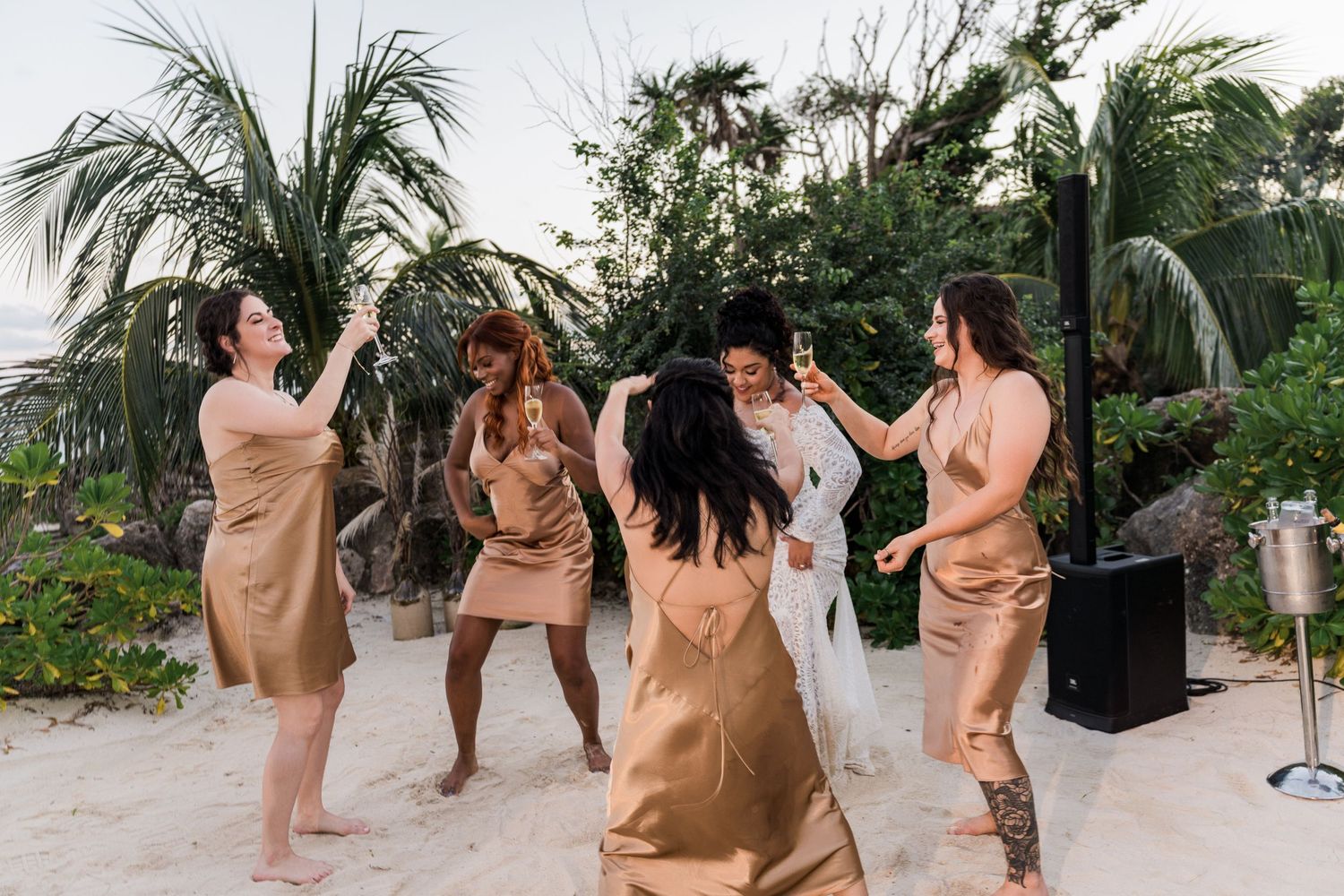 Xcaret Arte Wedding - Bridesmaids in gold satin dresses dance with bride on sandy beach surrounded by tropical palm trees.
