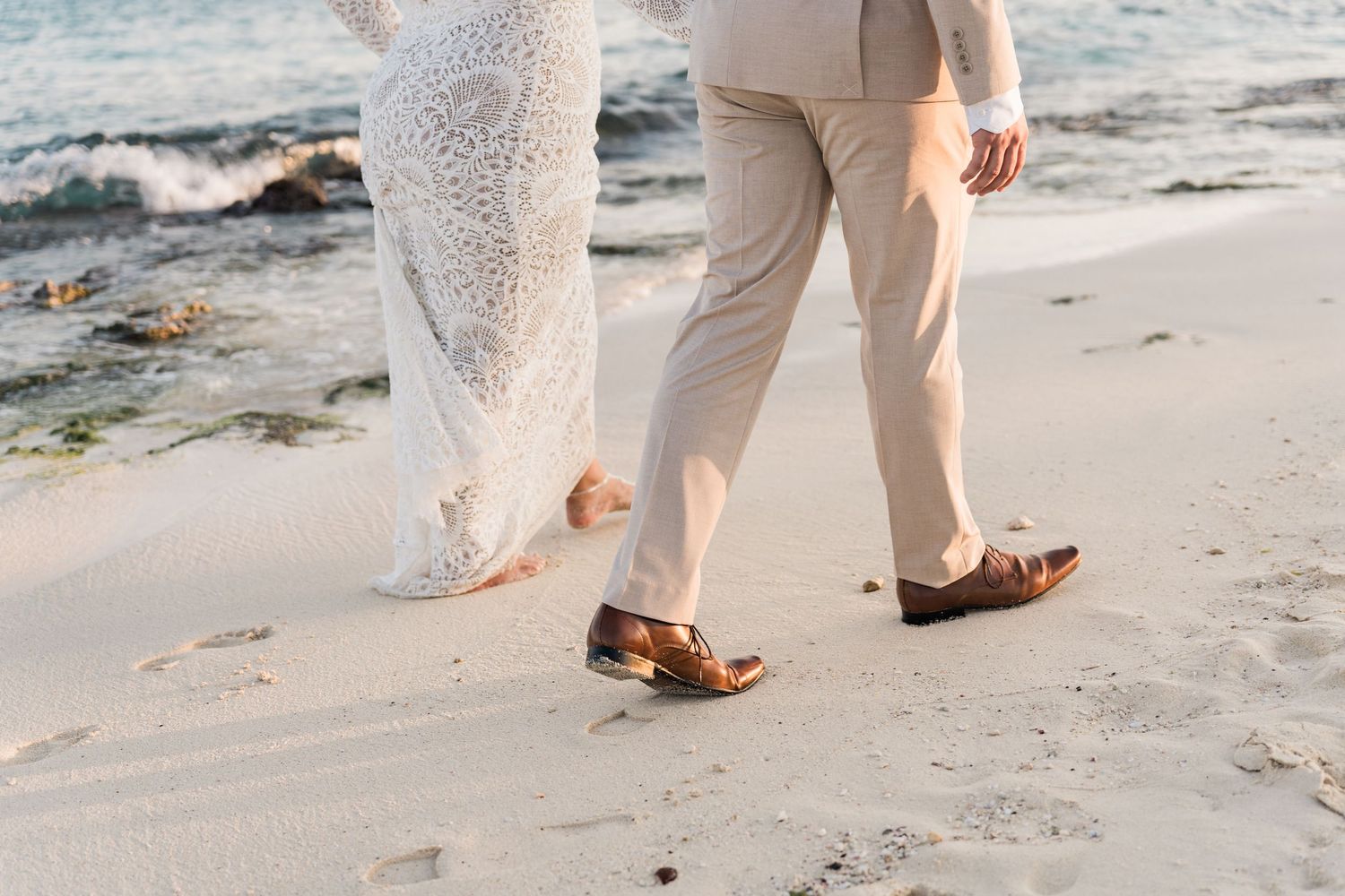 Xcaret Arte Wedding - Bride and groom walking barefoot on sandy beach, wedding dress and tan suit, ocean waves in background.