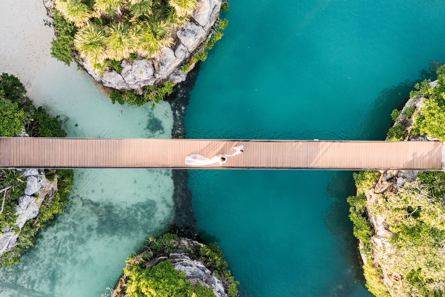 Xcaret Arte Wedding - Aerial view of a wooden suspension bridge over turquoise water between rocky islands with lush green trees.