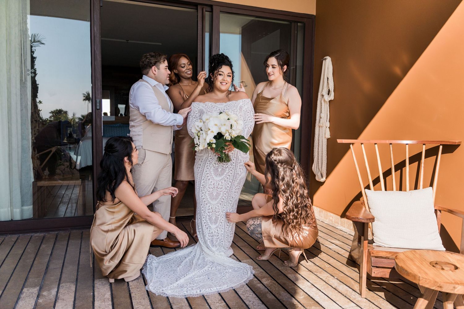 Xcaret Arte Wedding - Bride in white lace gown holds bouquet surrounded by bridesmaids in tan dresses on a wooden deck.