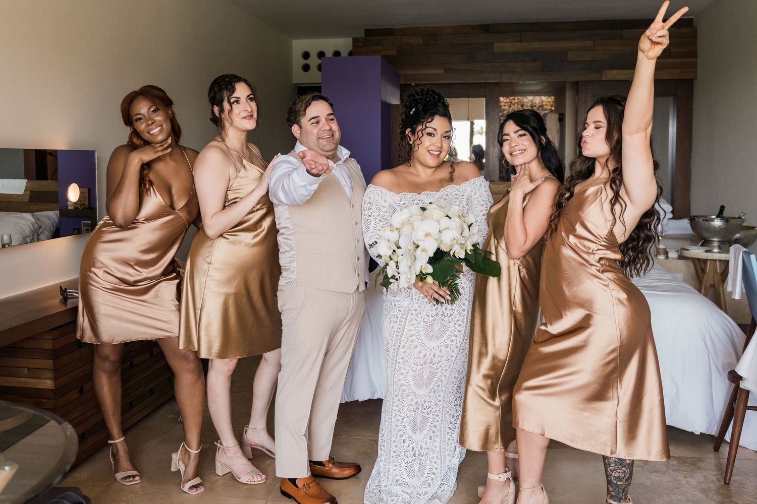 Xcaret Arte Wedding - Bride in lace gown holds white bouquet surrounded by bridesmaids in gold satin dresses celebrating indoors.