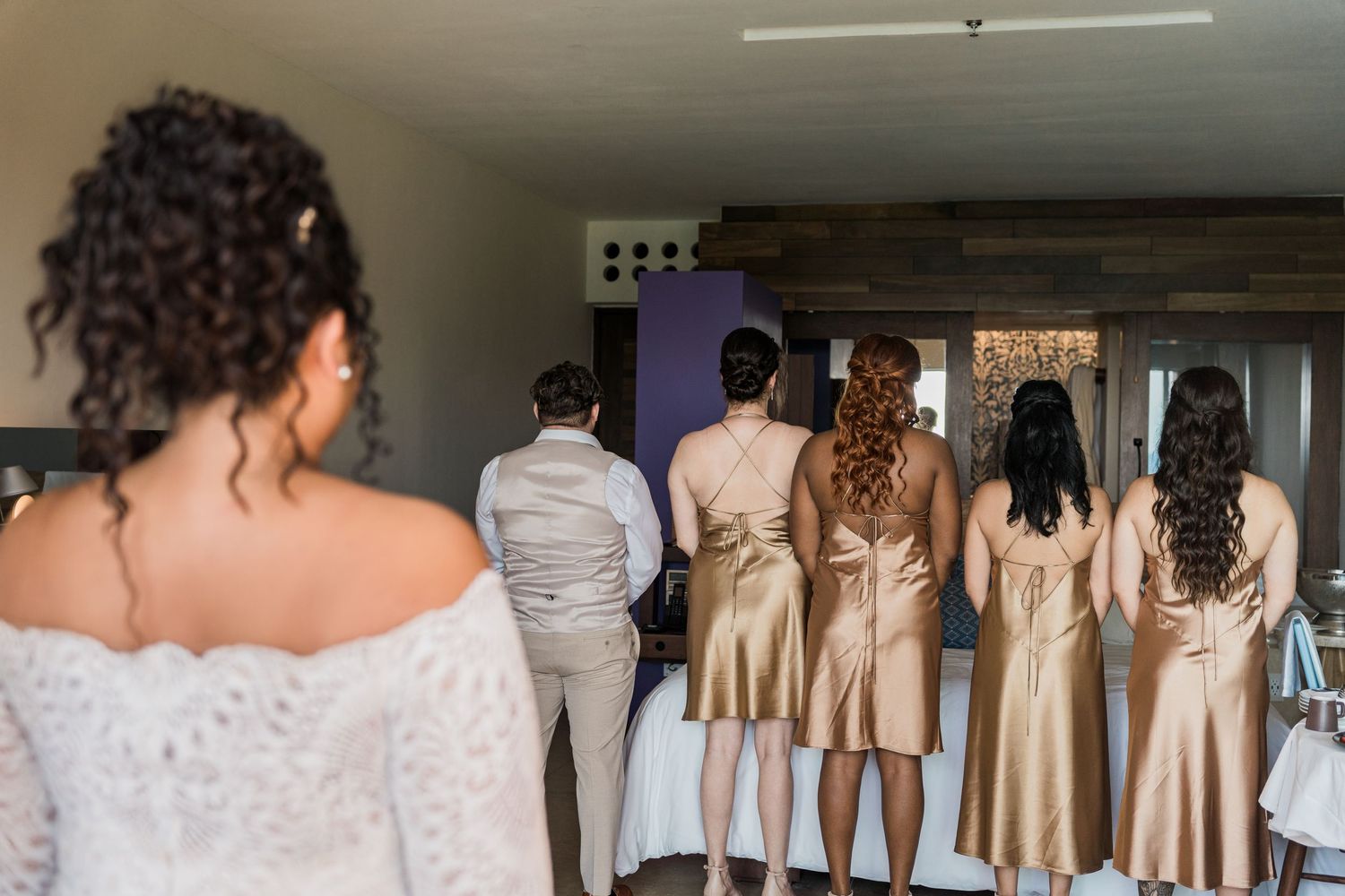 Xcaret Arte Wedding - Bride approaches bridesmaids in gold dresses standing with groomsman during wedding preparation indoors.