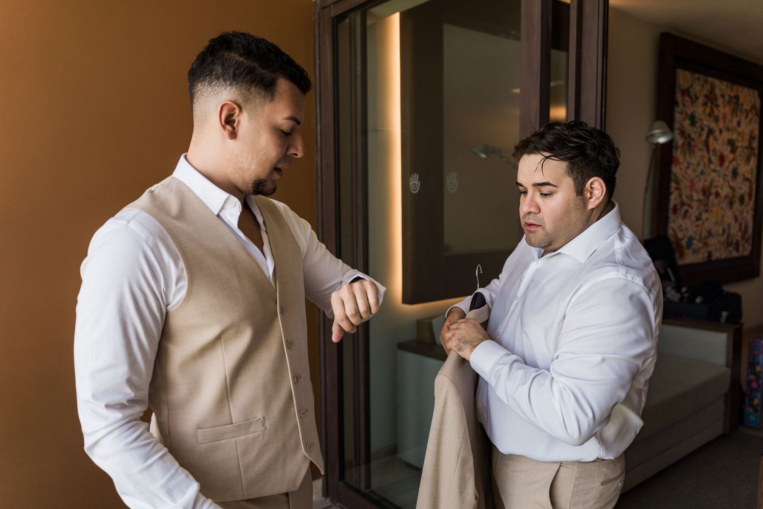 Xcaret Arte Wedding - Two groomsmen in white shirts and tan suits help each other get dressed before a wedding ceremony.
