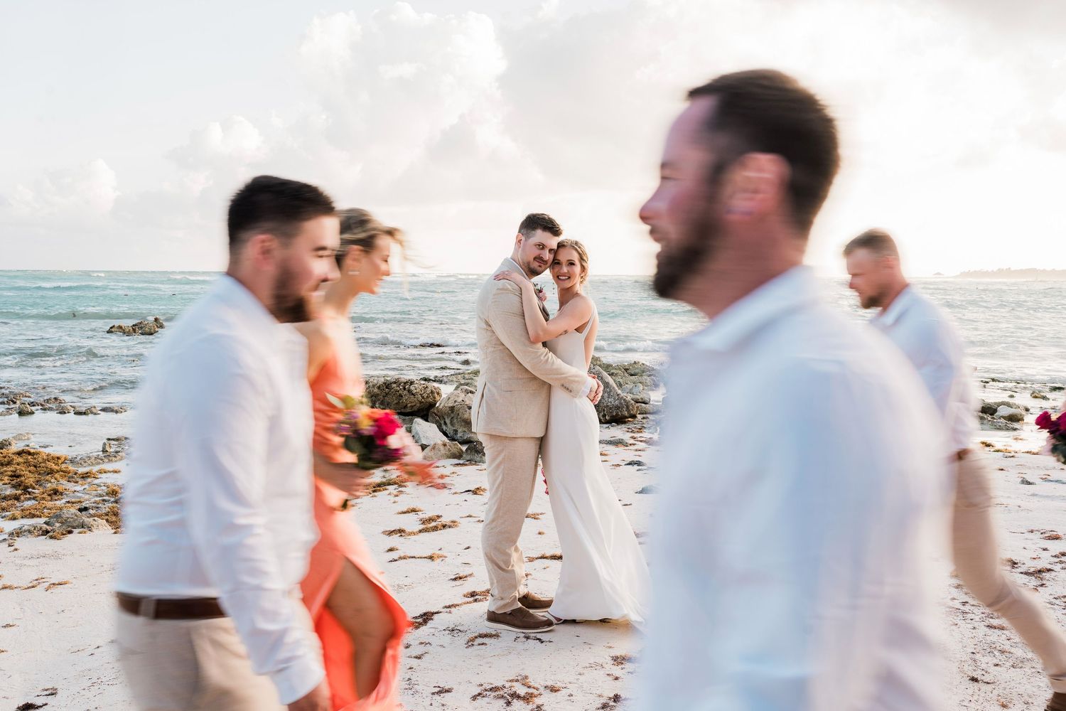 Bride and groom embrace on a tropical beach wedding ceremony with guests in white and coral attire by the ocean.