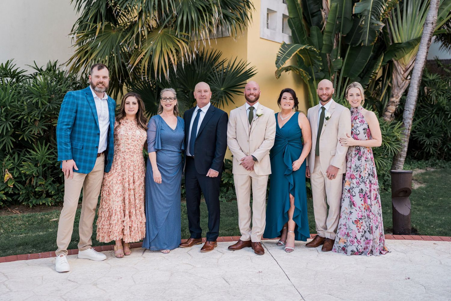 Group of eight adults posing together outdoors at a tropical resort, dressed in formal and semi-formal attire.