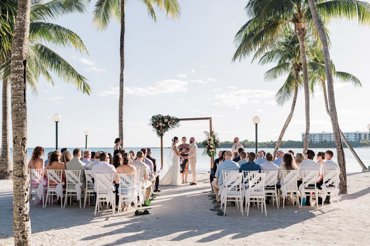 Outdoor beach wedding ceremony with guests seated on white chairs under palm trees near the ocean.