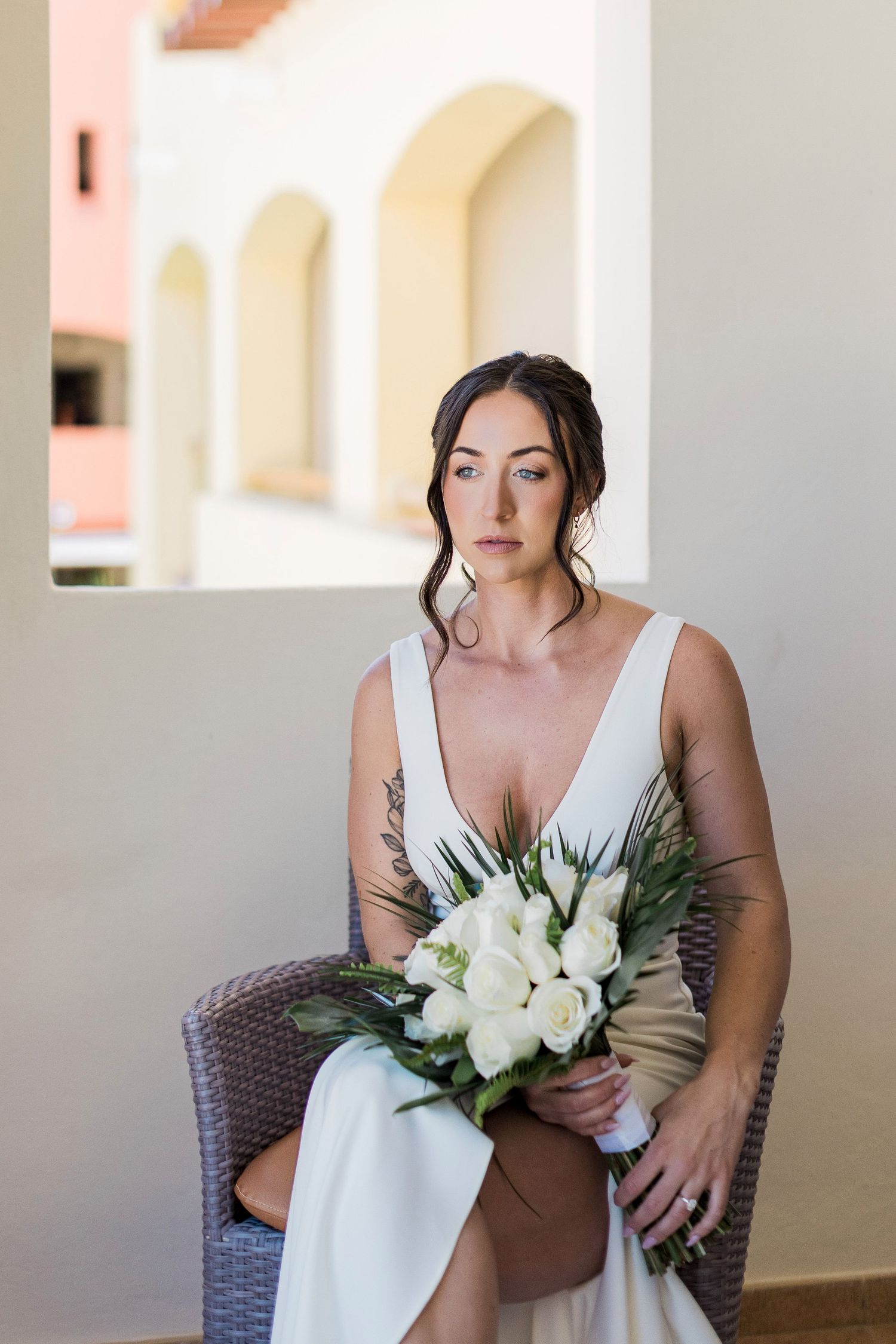 Bride in white gown holding white rose bouquet seated in wicker chair with arched architecture background.