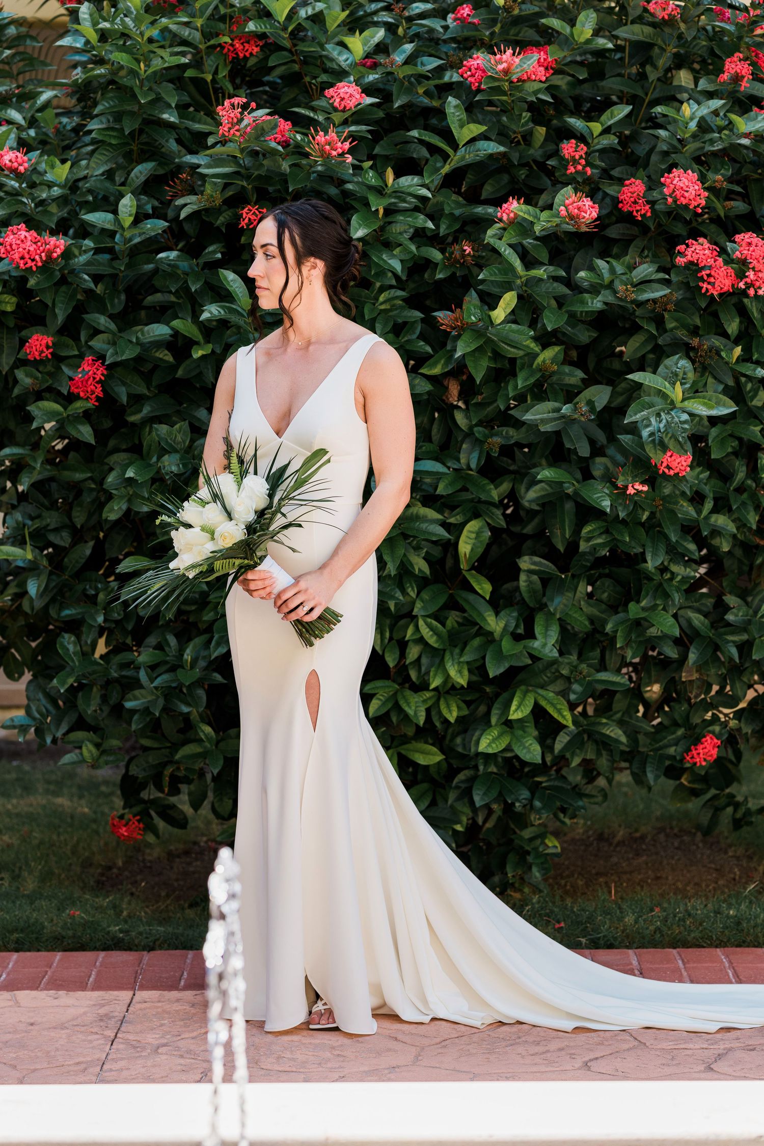 Bride in elegant white gown holding bouquet stands before lush green shrub with red flowers.