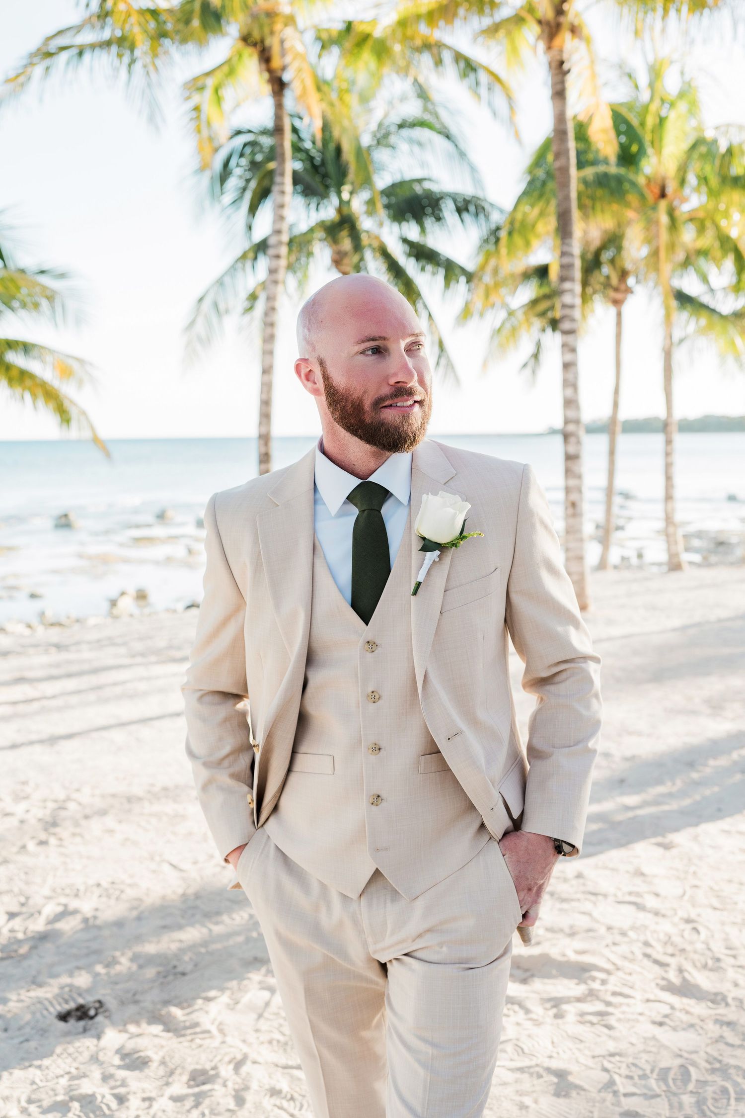 Groom in beige three-piece suit with dark green tie and white boutonniere poses on sandy beach with palm trees.