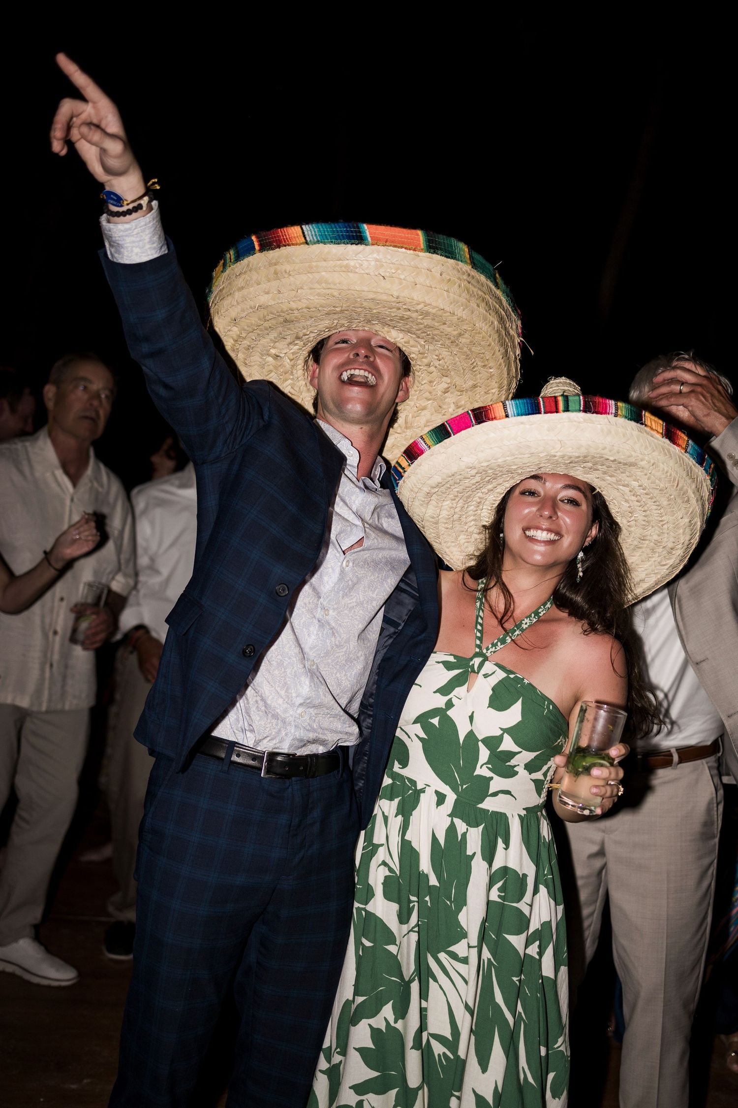 Couple wearing colorful sombreros celebrating at a festive outdoor party, man pointing up while woman smiles holding a drink.