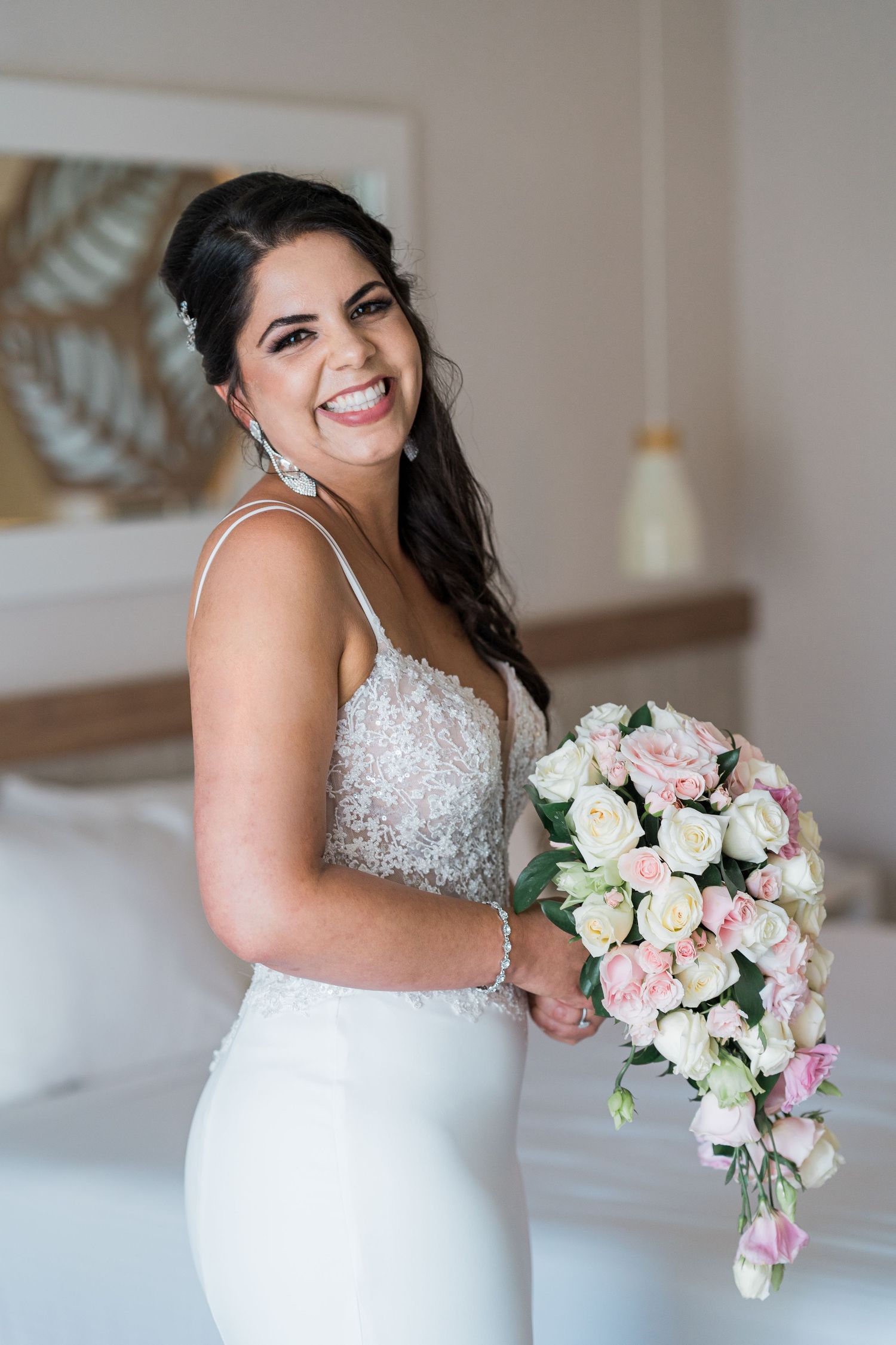 Smiling bride in a beaded white gown holding a cascading pink and white rose bouquet in a bright hotel room.