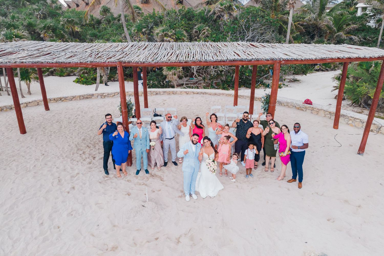 Aerial view of wedding party and guests posing together under a rustic beach palapa structure.