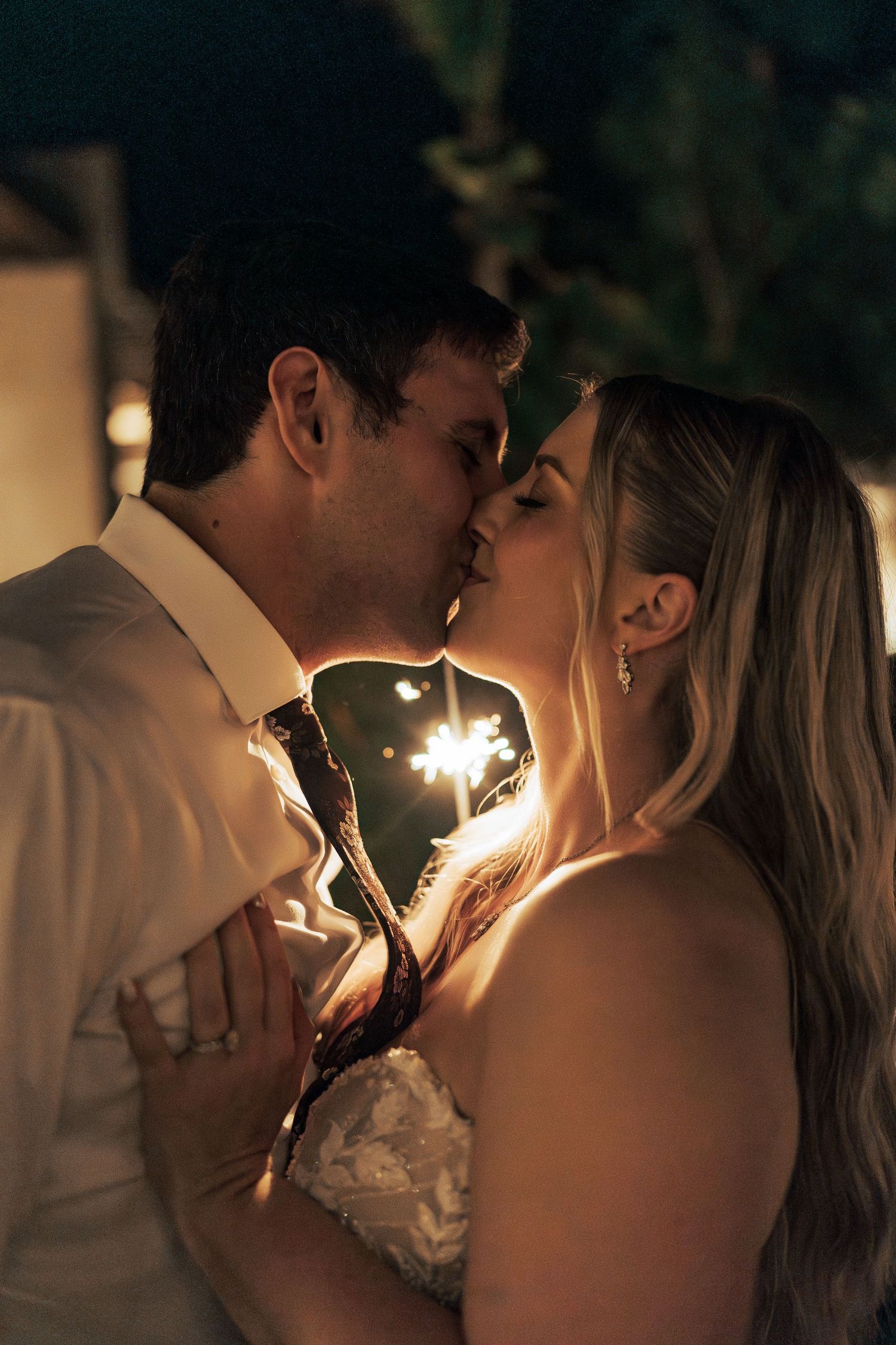 Bride and groom sharing a romantic kiss with sparklers glowing at night.