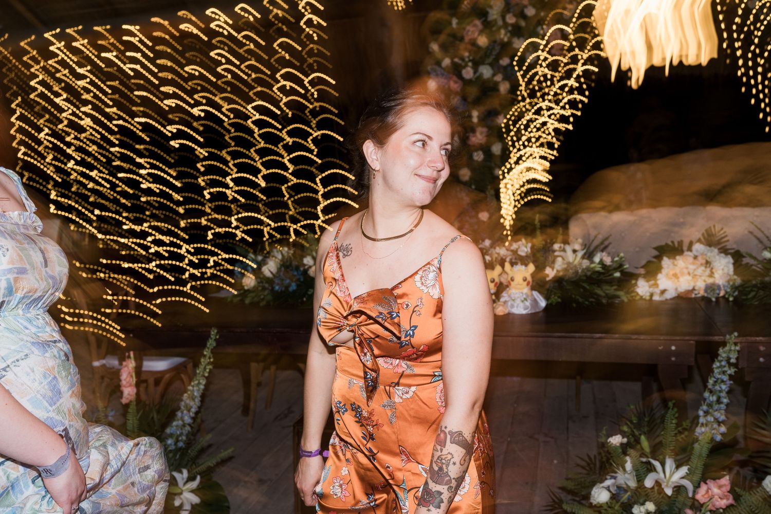 Woman in orange floral dress smiling at elegant indoor wedding reception with twinkling lights.