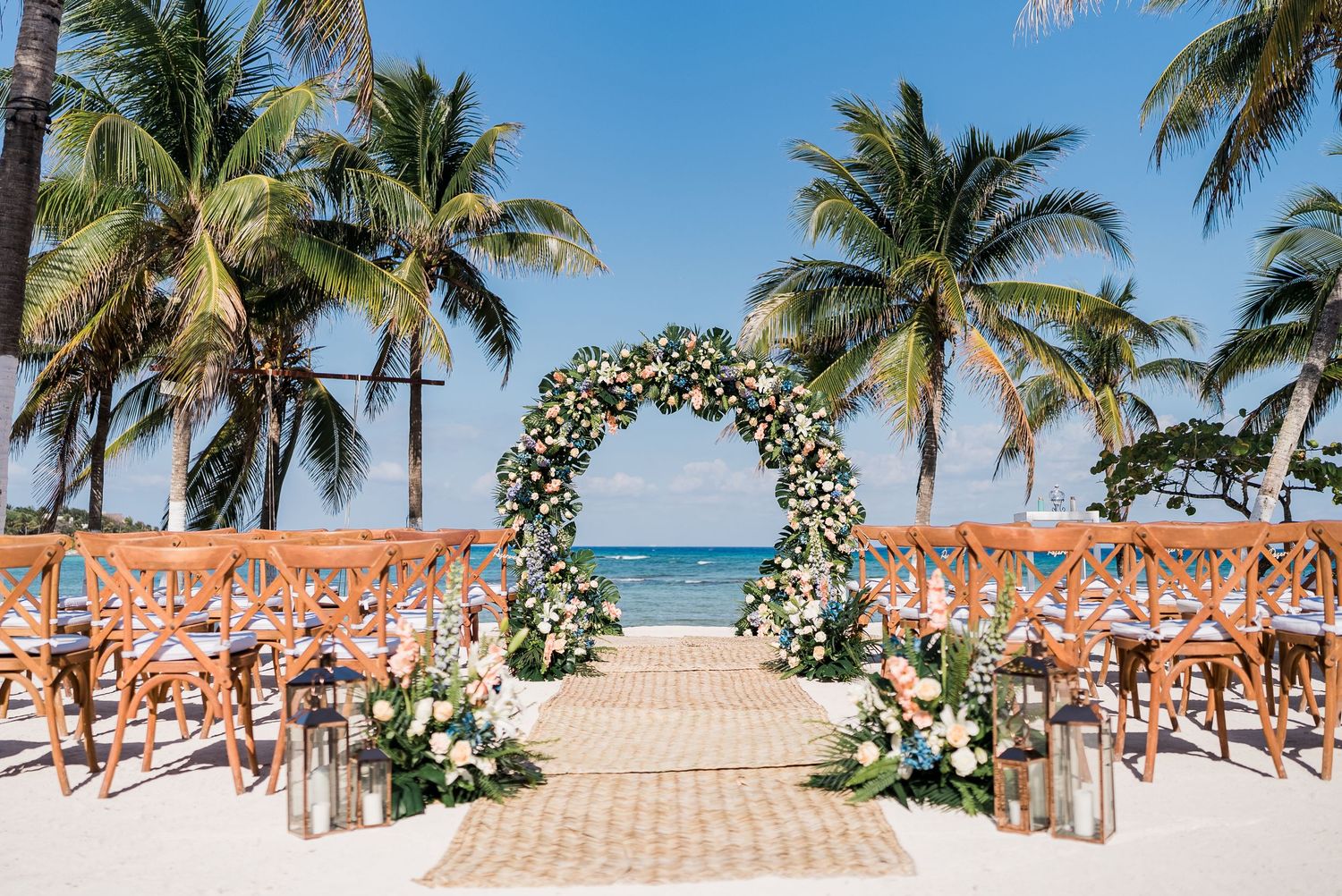Elegant floral arch wedding ceremony setup on a tropical beach with palm trees and ocean views.