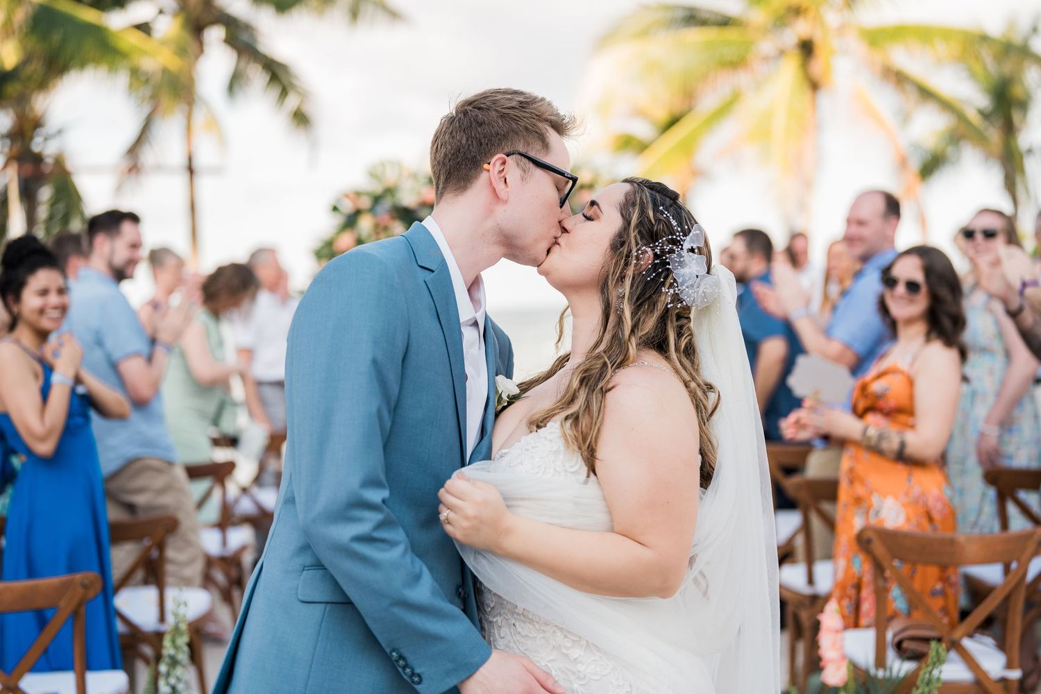 Bride and groom share romantic kiss while guests applaud at outdoor beach wedding.
