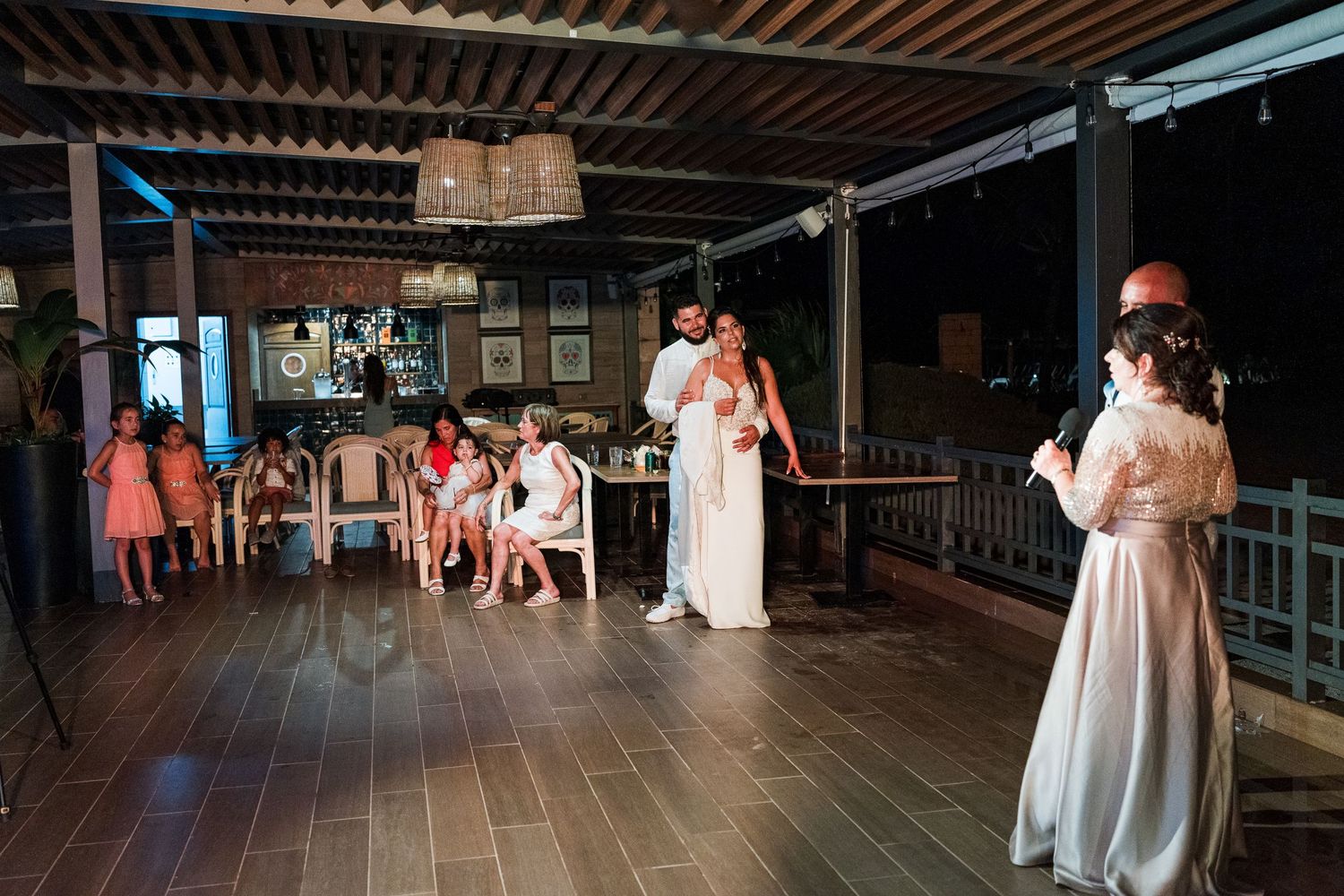 Bride and groom standing together on a wooden dance floor as a guest performs a wedding speech.
