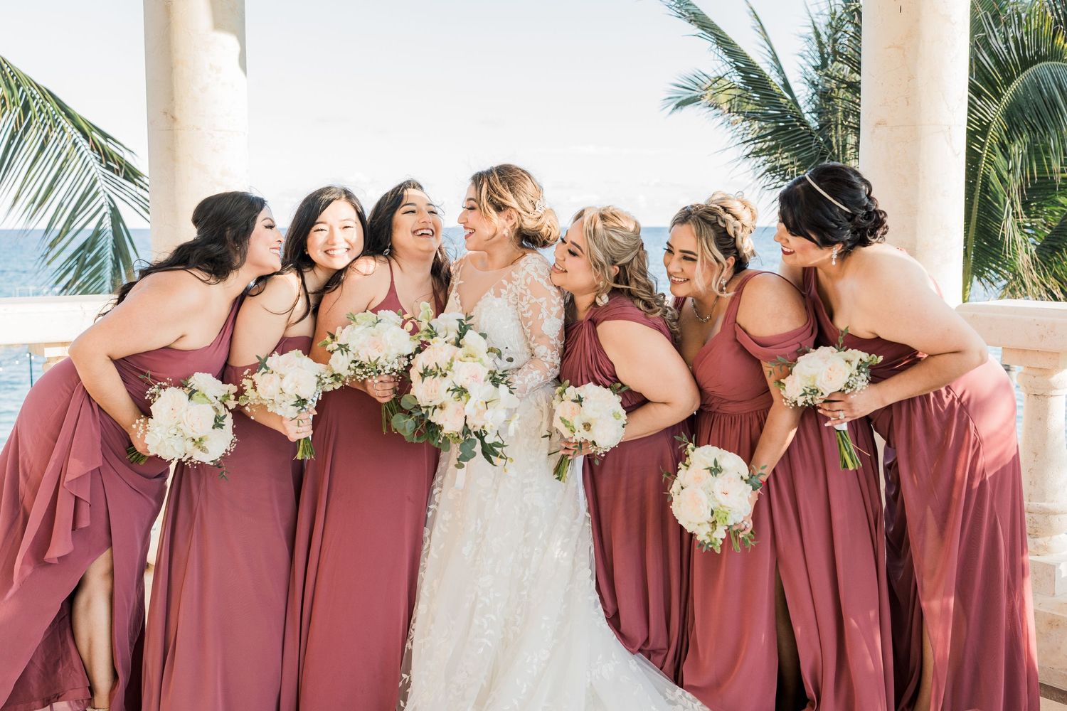 Bride in lace gown laughing with six bridesmaids in dusty rose dresses holding white bouquets at oceanside wedding.