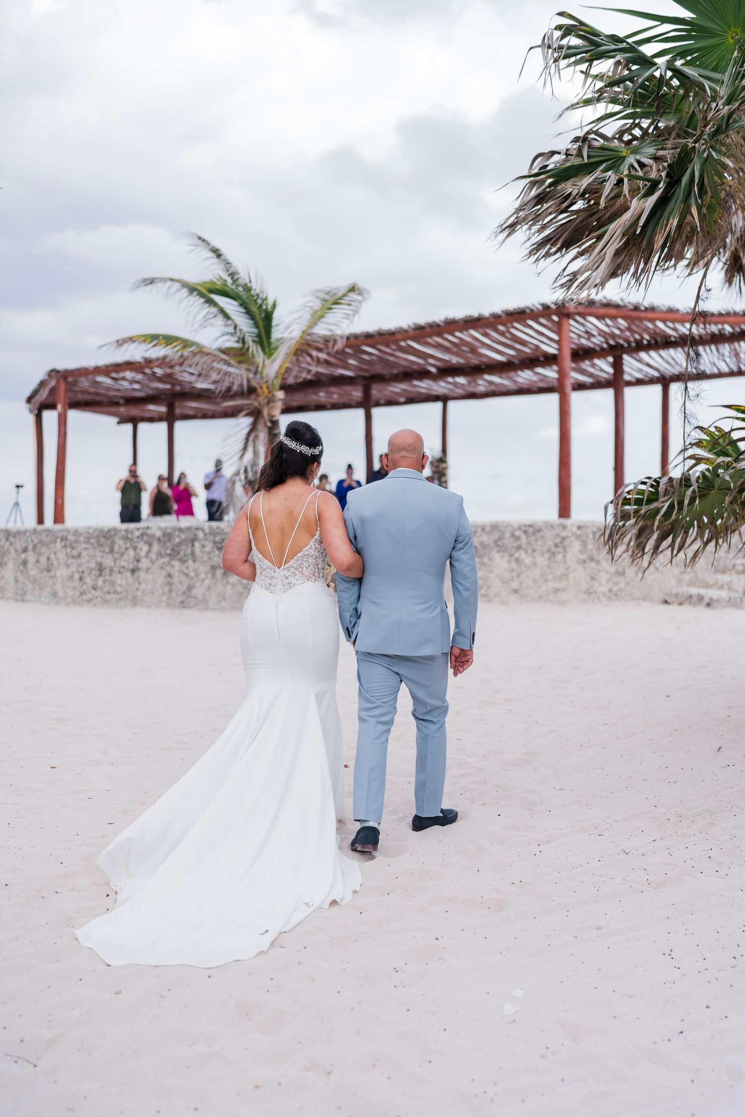 Bride in backless white gown walks with escort toward beach wedding pergola on white sand.