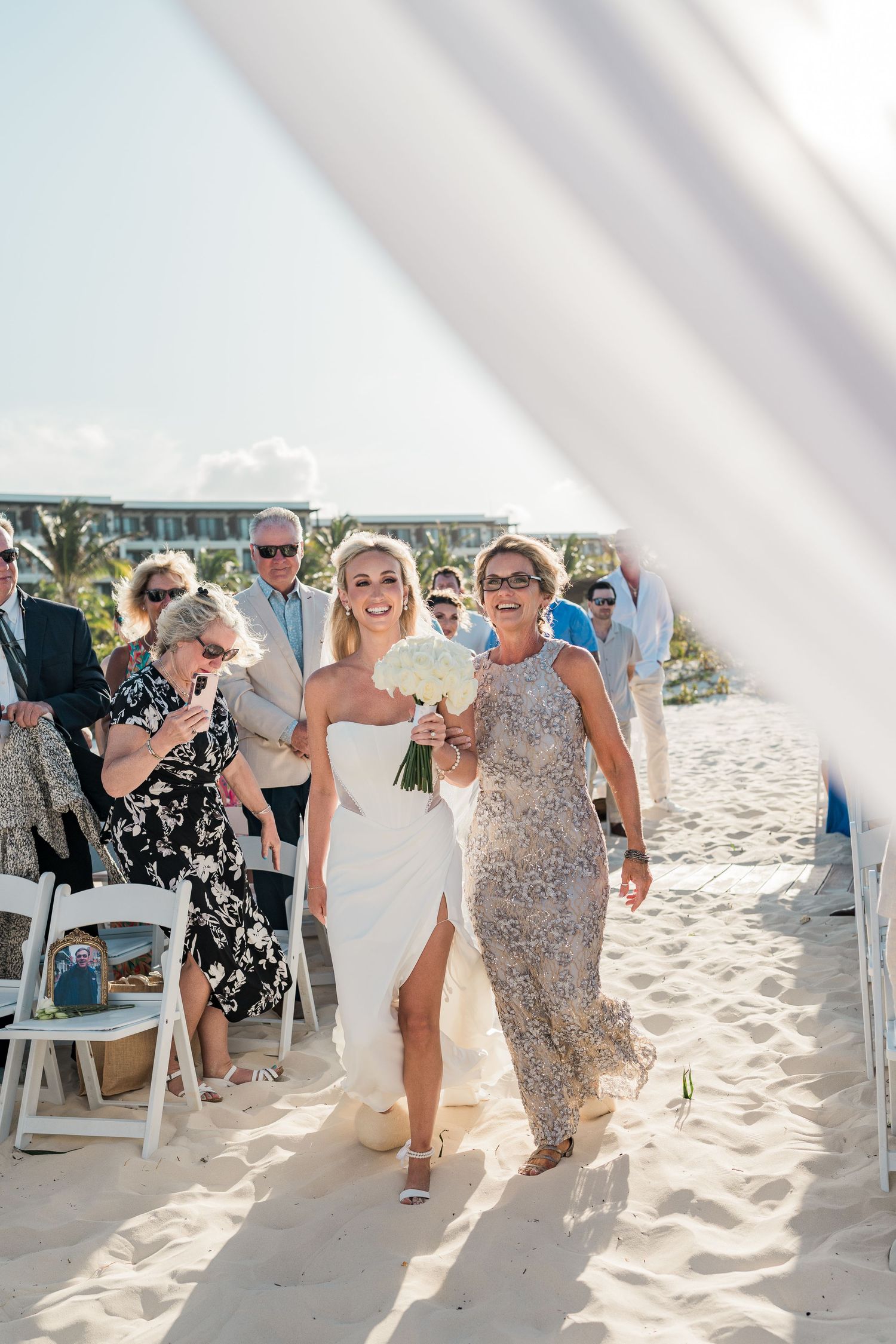 Bride walks down sandy beach aisle with her mother, both holding white bouquets, guests seated on white chairs.