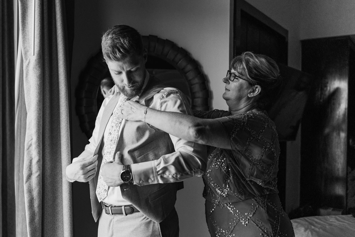 Black and white photo of groom being helped into suit jacket by older woman before wedding.
