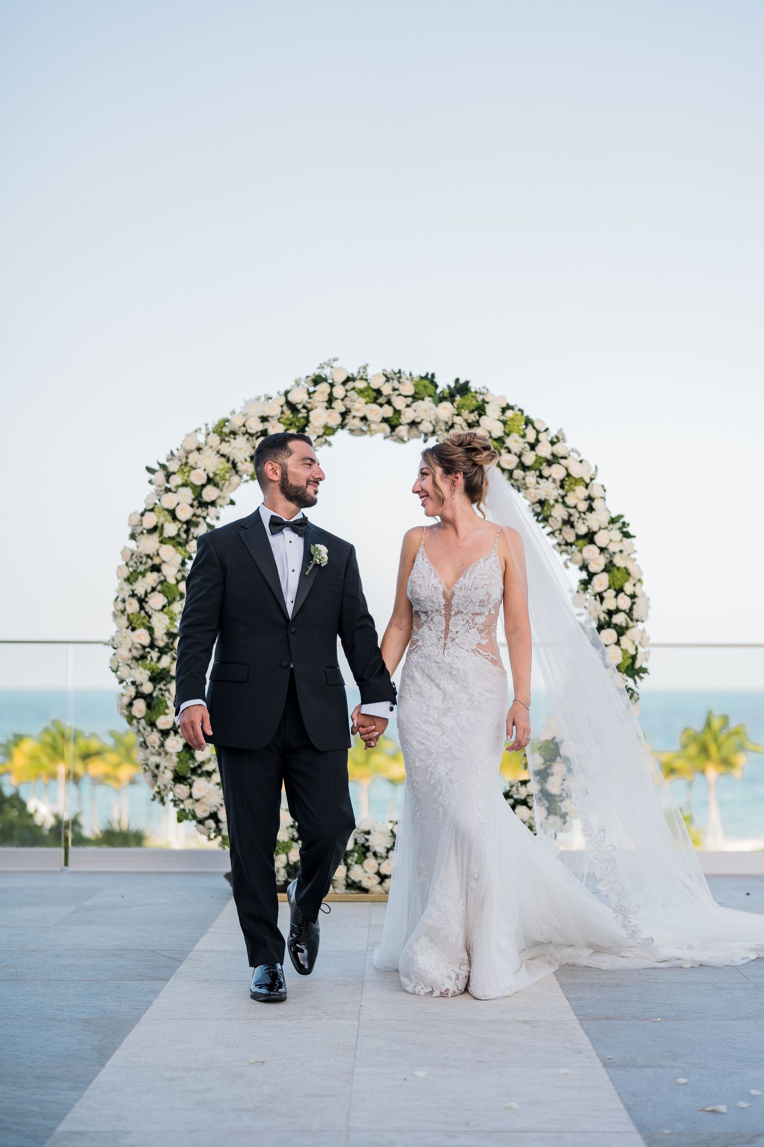 Newlyweds walk hand in hand from white floral arch at romantic beachside wedding ceremony.