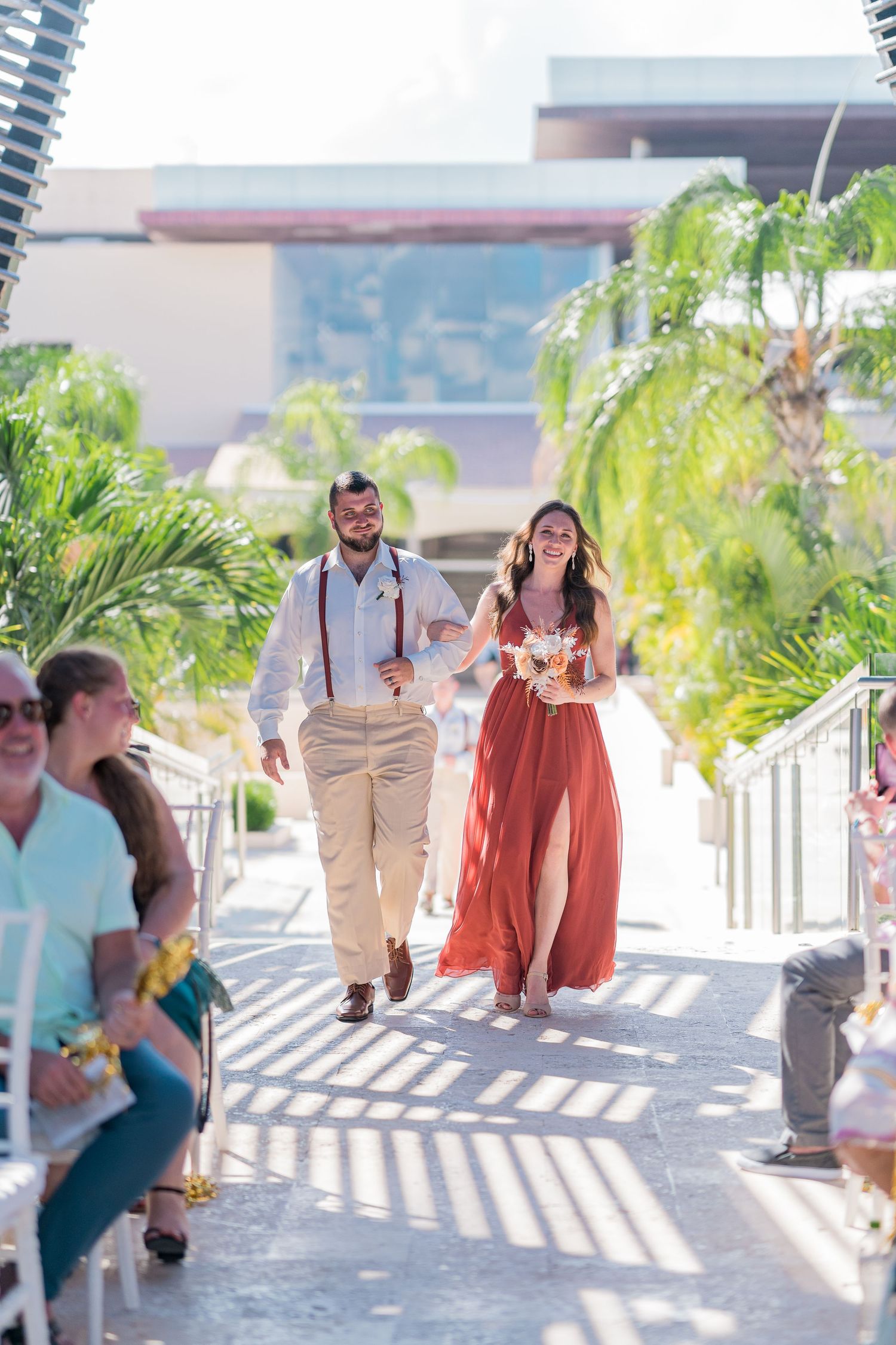Groomsman in suspenders escorts bridesmaid in red dress down sunny outdoor wedding aisle.