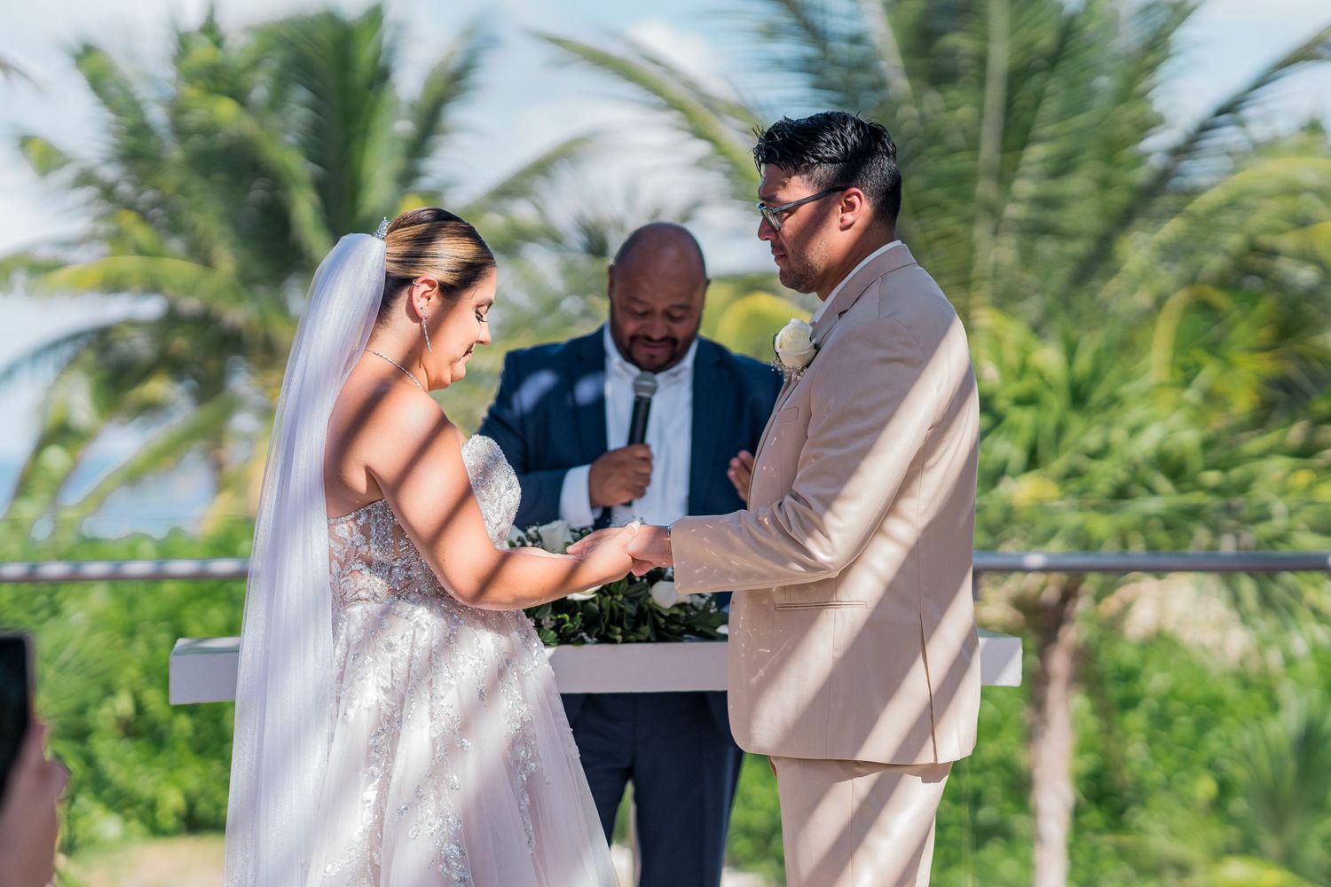 Groom places ring on brides finger during outdoor beach wedding ceremony with officiant.