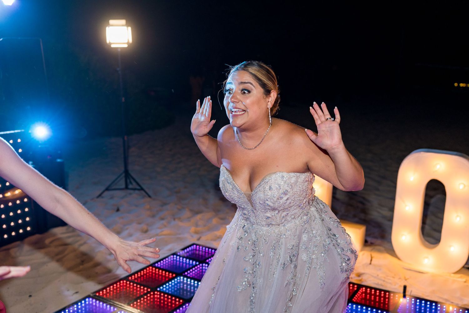 Bride in strapless wedding gown dancing joyfully on colorful LED dance floor at night.