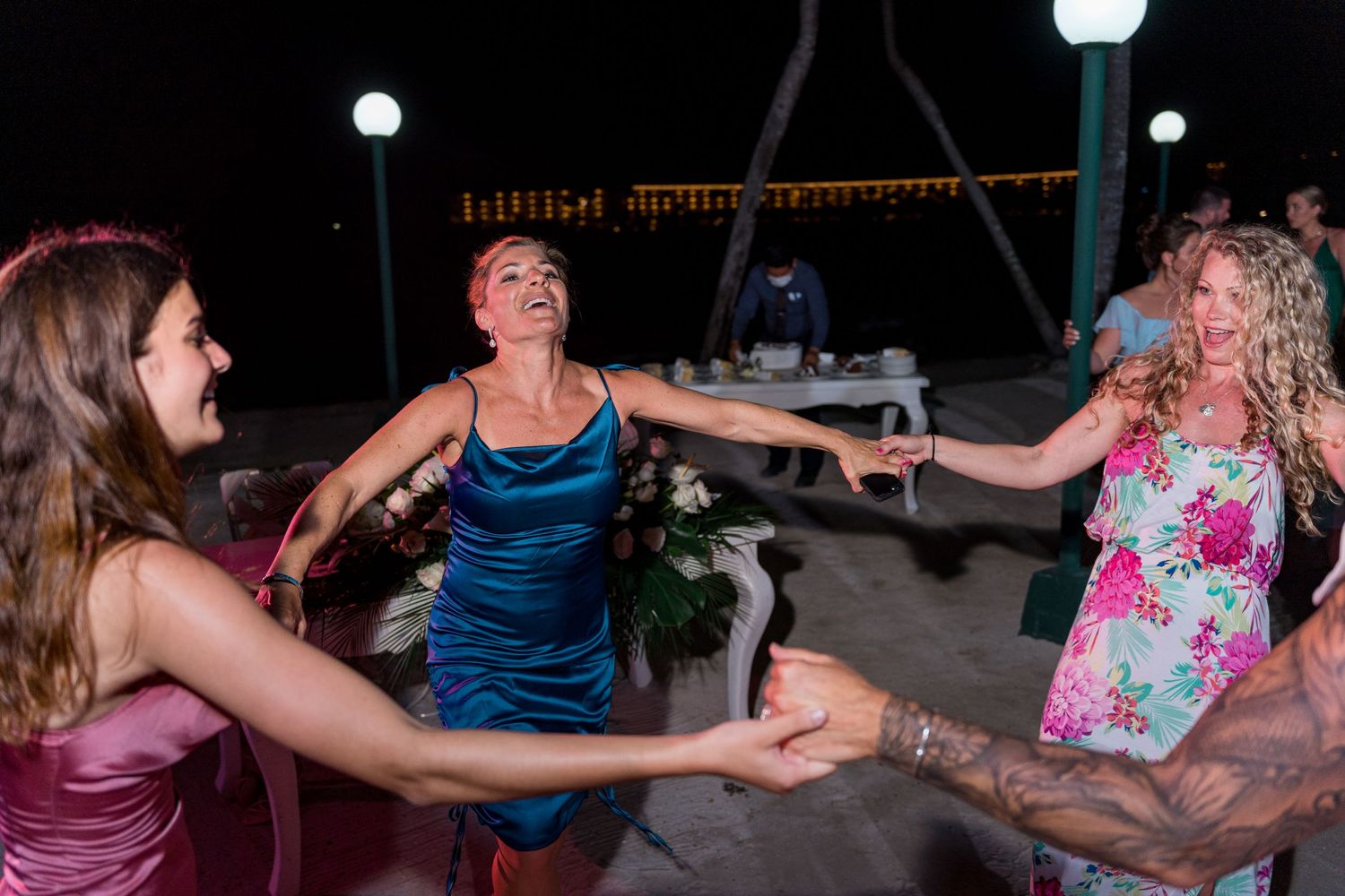 Women hold hands and dance together joyfully at an outdoor nighttime beach wedding reception.