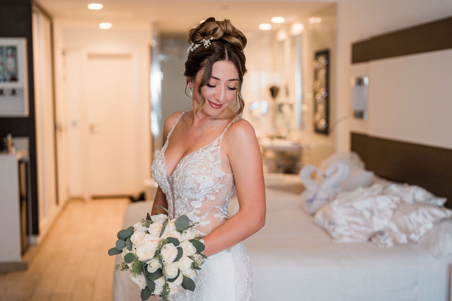 Bride in lace gown looking down at white rose and eucalyptus bouquet.