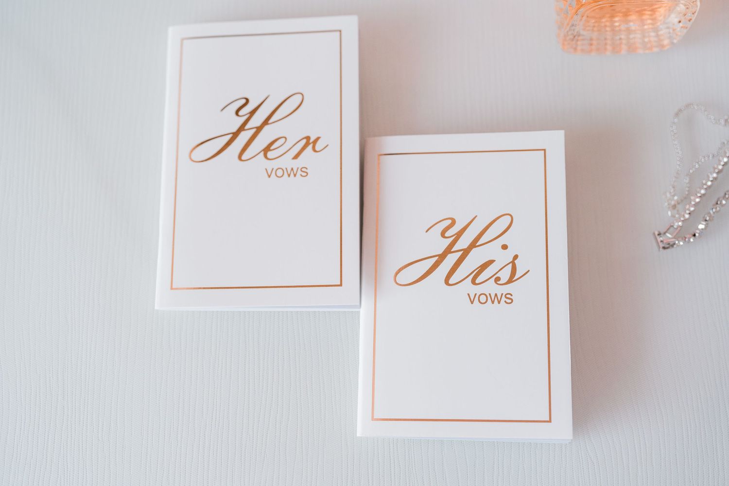 Two elegant white and rose gold foil wedding vow books labeled Her Vows and His Vows on a white surface.