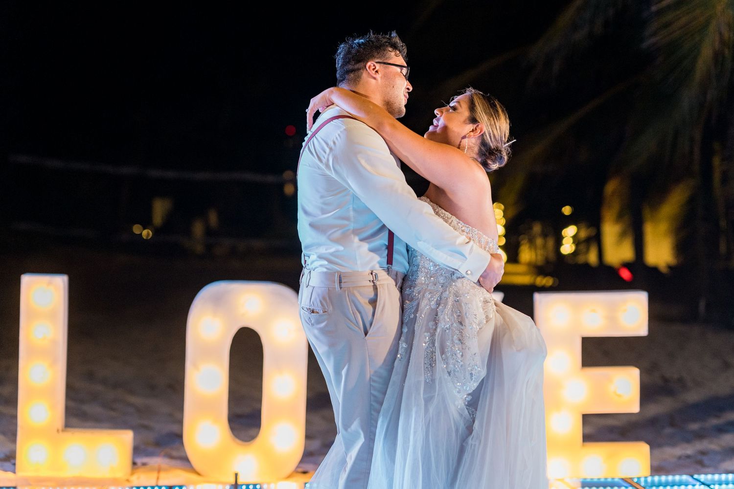 Couple sharing romantic kiss in front of illuminated LOVE letters on beach at night wedding.