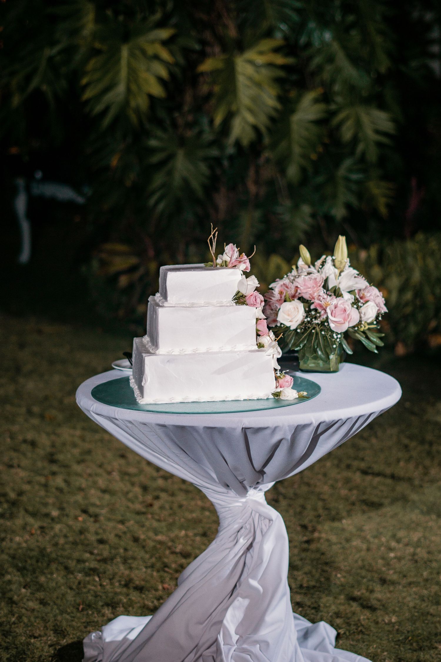 Elegant three-tier white square wedding cake with pink roses on a draped cocktail table outdoors.